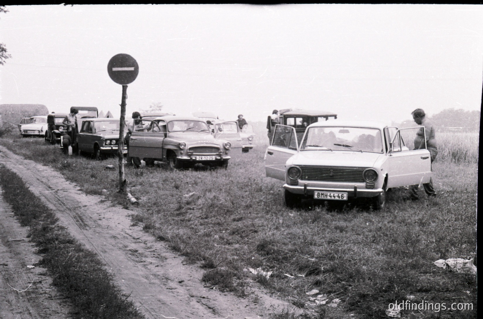 Vintage black-and-white roadside scene with 1970s-era cars parked on grass beside a rural road. Soviet-era vehicles dominate, including a prominent white Lada and a dark sedan. A no-entry traffic sign stands near the road. People in casual attire stand or lean against vehicles, suggesting a social gathering or roadside stop. Overcast skies and muddy ground enhance the nostalgic atmosphere.