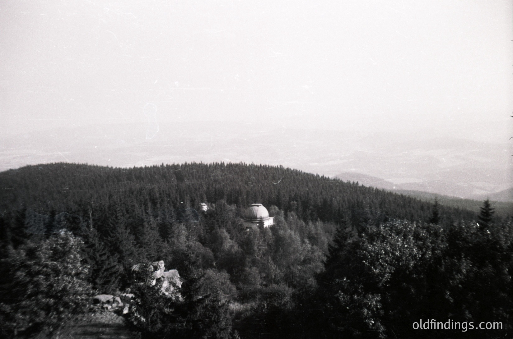 Vintage black-and-white aerial view of a domed structure nestled in a forested hillside, likely a Soviet-era observation tower or radio station. Dense pine forest surrounds the base, with expansive plains visible in the distance.