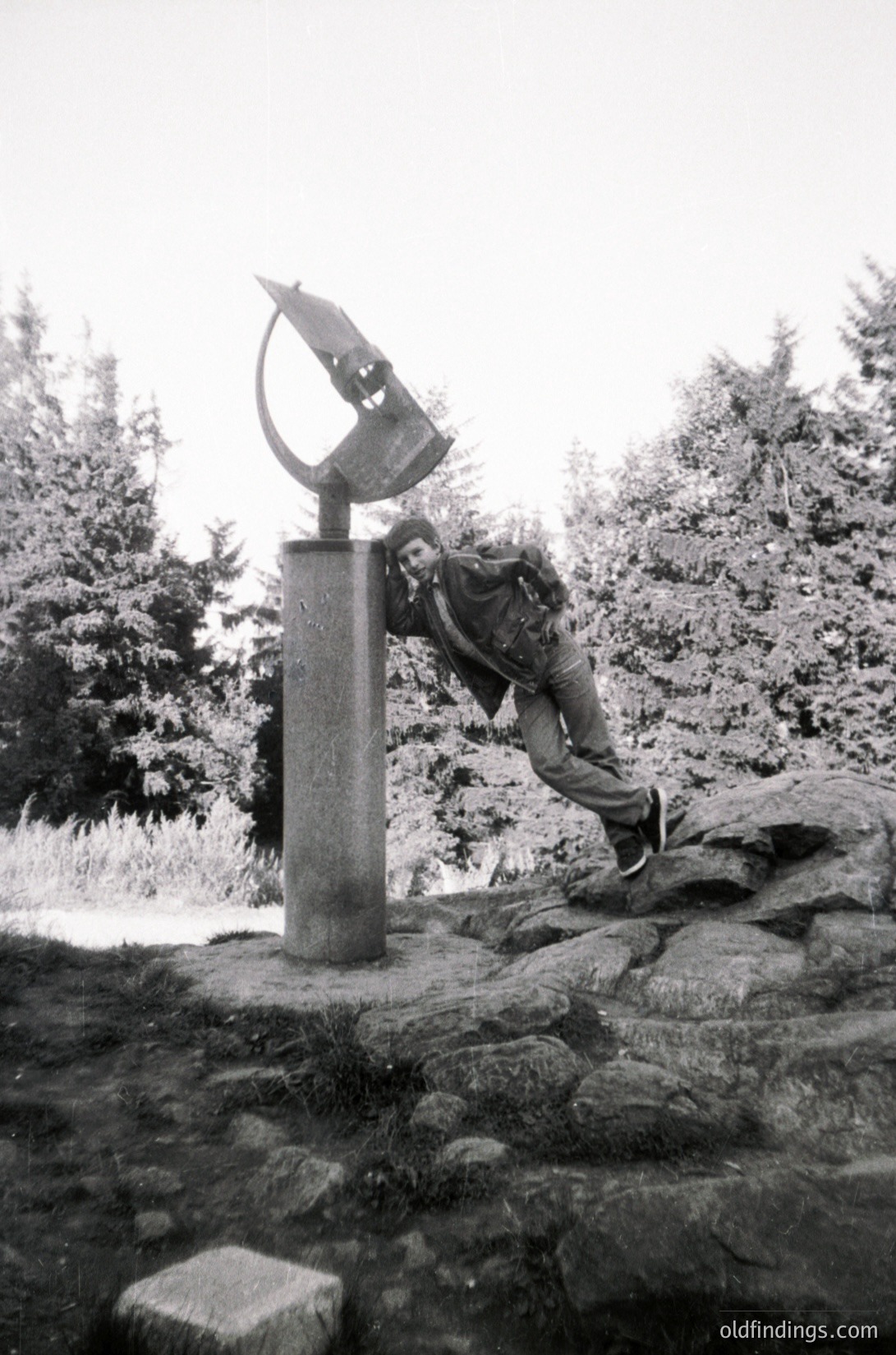 Black-and-white photo of a person adjusting a large, abstract sundial sculpture atop a stone pedestal in a forested area. The sculpture resembles a stylized bird or arrow. Mid-20th century outdoor art installation, likely or .