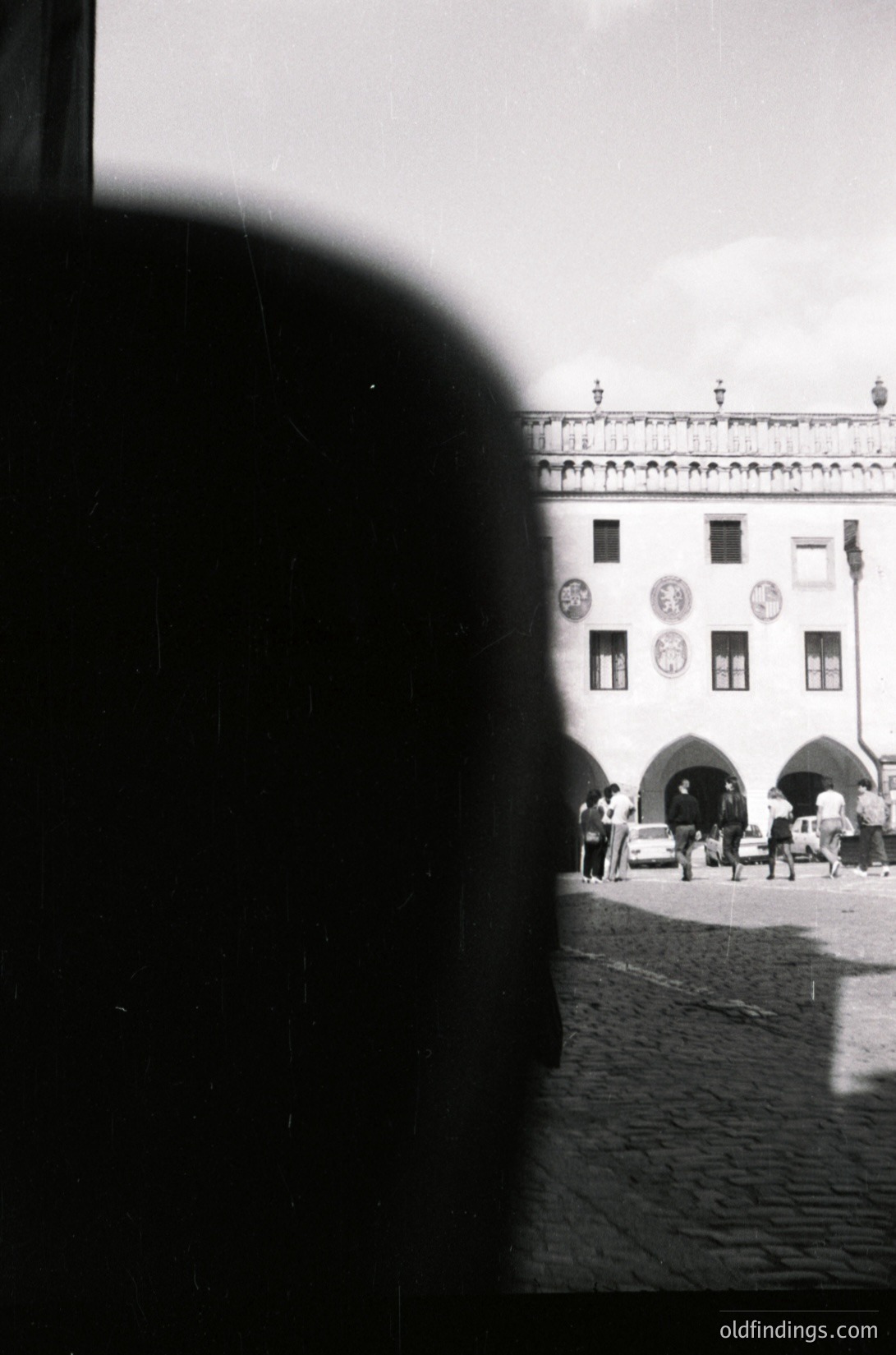 Black-and-white shot of a cobblestone plaza framed by a blurred silhouette. Prominent neoclassical building with arched entrance, decorative clock faces, and balustrade. Mid-20th century European urban scene.