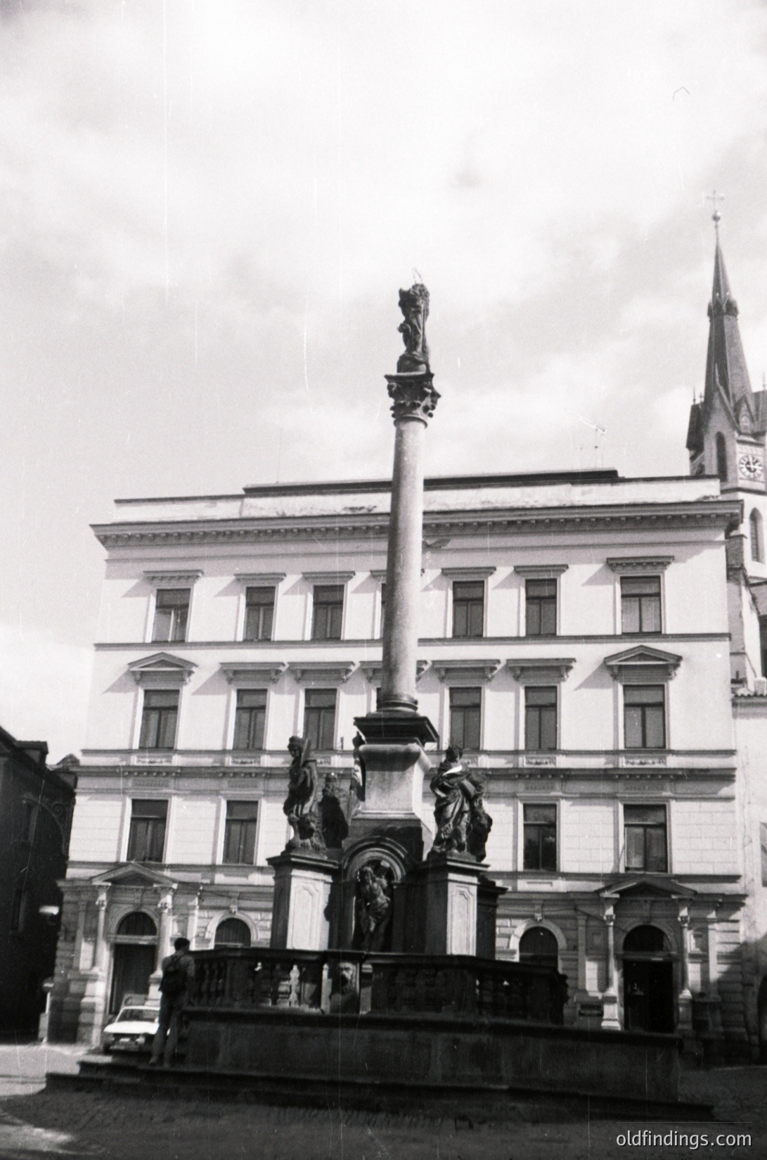 Neoclassical monument with tall column and sculptural reliefs in front of a multi-story institutional building, likely from the early-to-mid 20th century. The column features a central figure atop, flanked by decorative carvings. Symmetrical architecture with arched doorways and rectangular windows. Overcast sky enhances dramatic contrast.