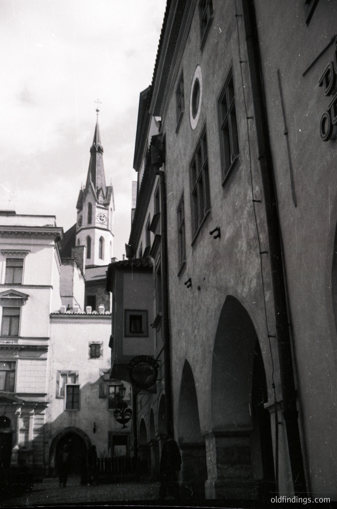 Black-and-white street scene featuring Gothic Revival architecture. Prominent church spire with ornate detailing and clock tower in background. Narrow alley flanked by aged stone buildings with arched doorways and small windows. Street sign visible, indicating a European urban setting. Likely mid-20th century.