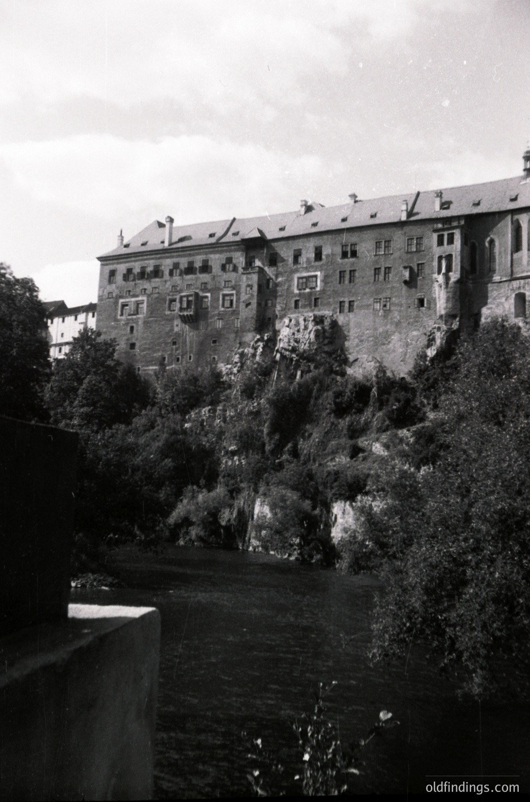Historic castle perched on rocky cliffs beside a river, featuring symmetrical brick architecture with numerous windows and chimneys. Likely European, mid-20th century.