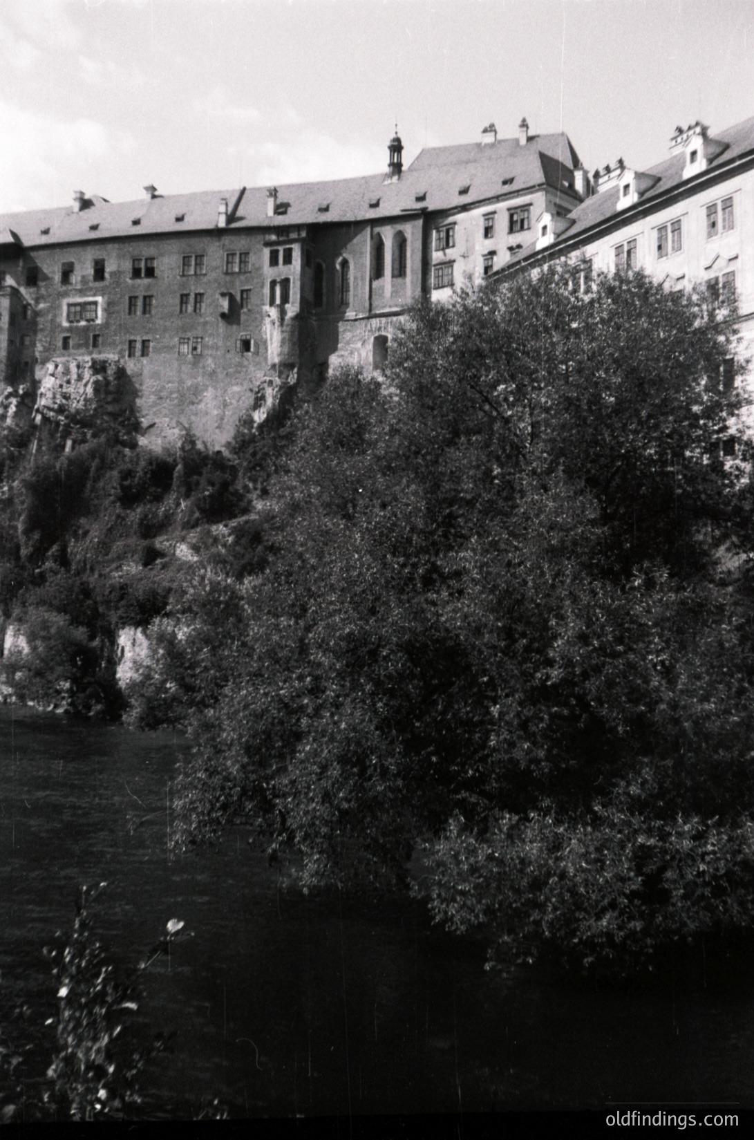 Historic stone fortress perched on rocky cliffs beside a river, mid-20th century. Symmetrical towers and battlements suggest European medieval or Renaissance architecture. Dense foliage partially obscures lower structures.