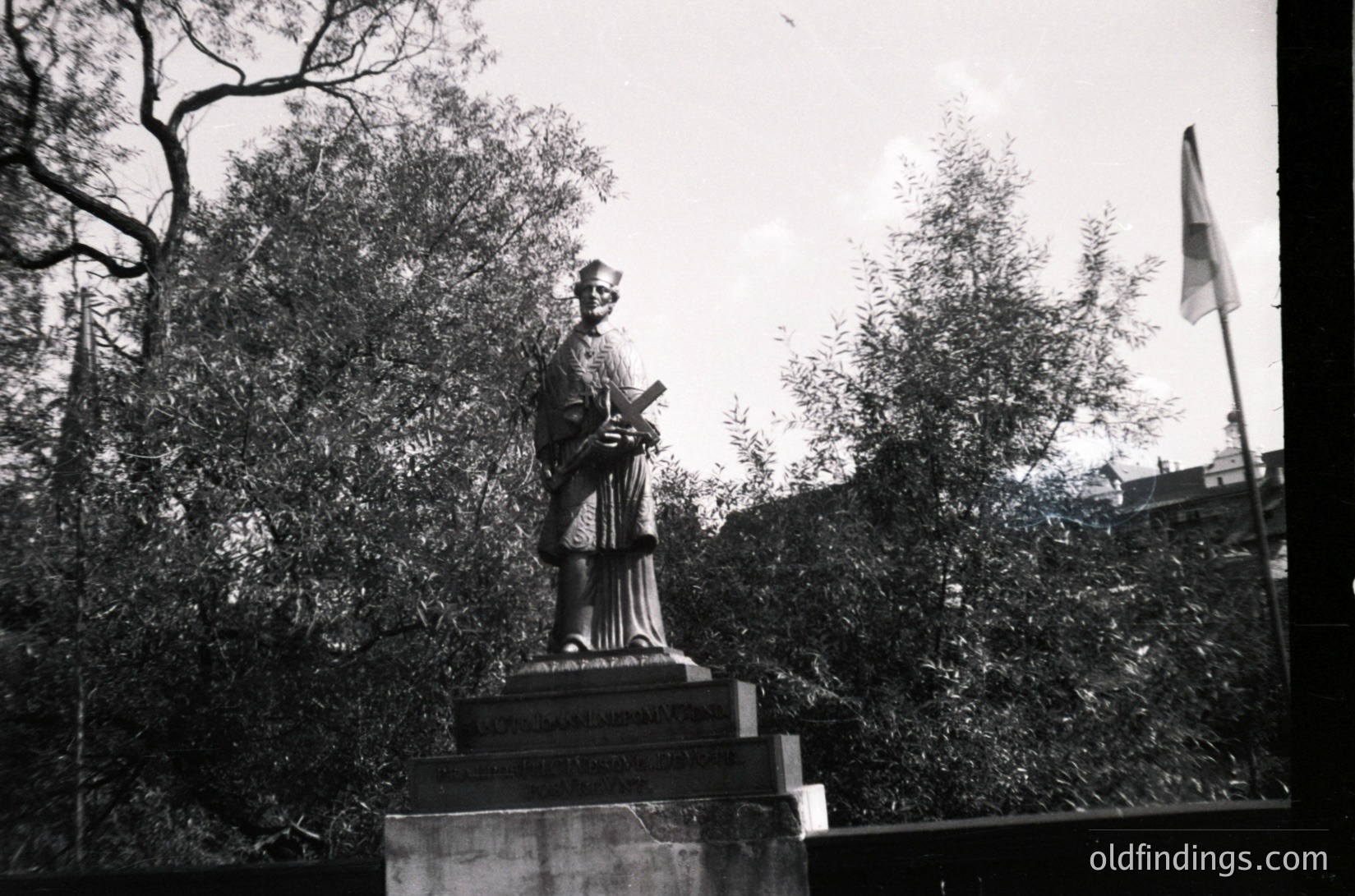 Statue of a robed figure holding a scroll, mounted on a pedestal with inscribed text, set in a landscaped park. Flagpole with a red-and-white striped flag in background. Black-and-white photo, likely –.