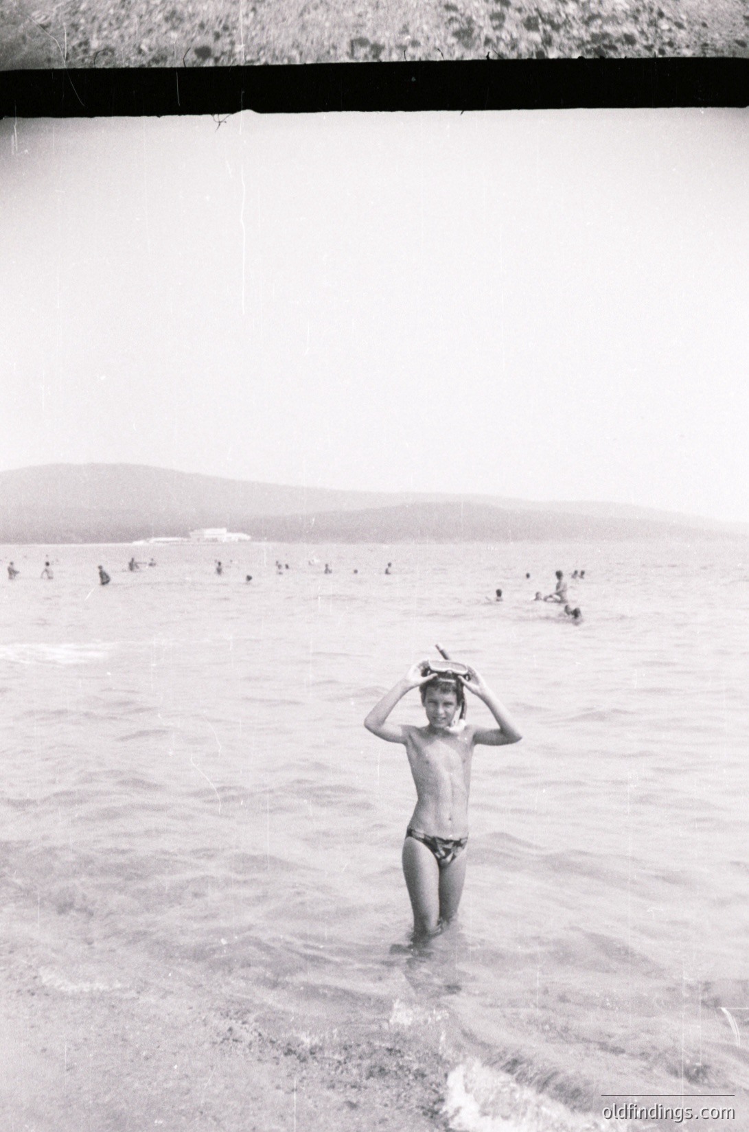 Mid-20th century black-and-white seaside scene: boy in swim trunks stands waist-deep in shallow water, adjusting a hat. Crowded beach with figures in water and on shore, framed by a hotel balcony.