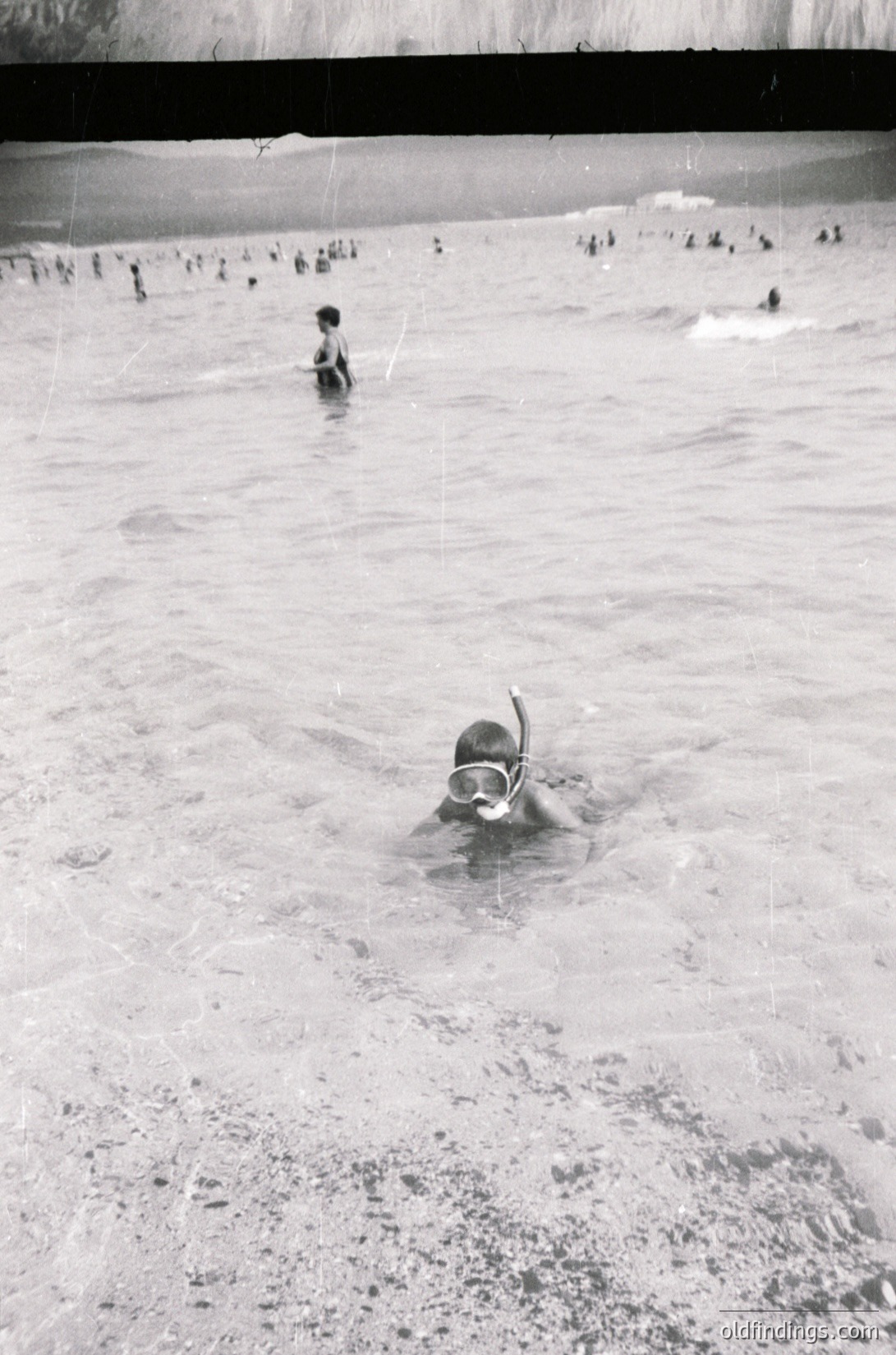 Black-and-white shot of a crowded outdoor swimming pool, likely from the 1950s–1970s. A child in a snorkel mask dives near shallow water, while others swim or wade in the background. Mid-century concrete pool design with visible tiled edges.