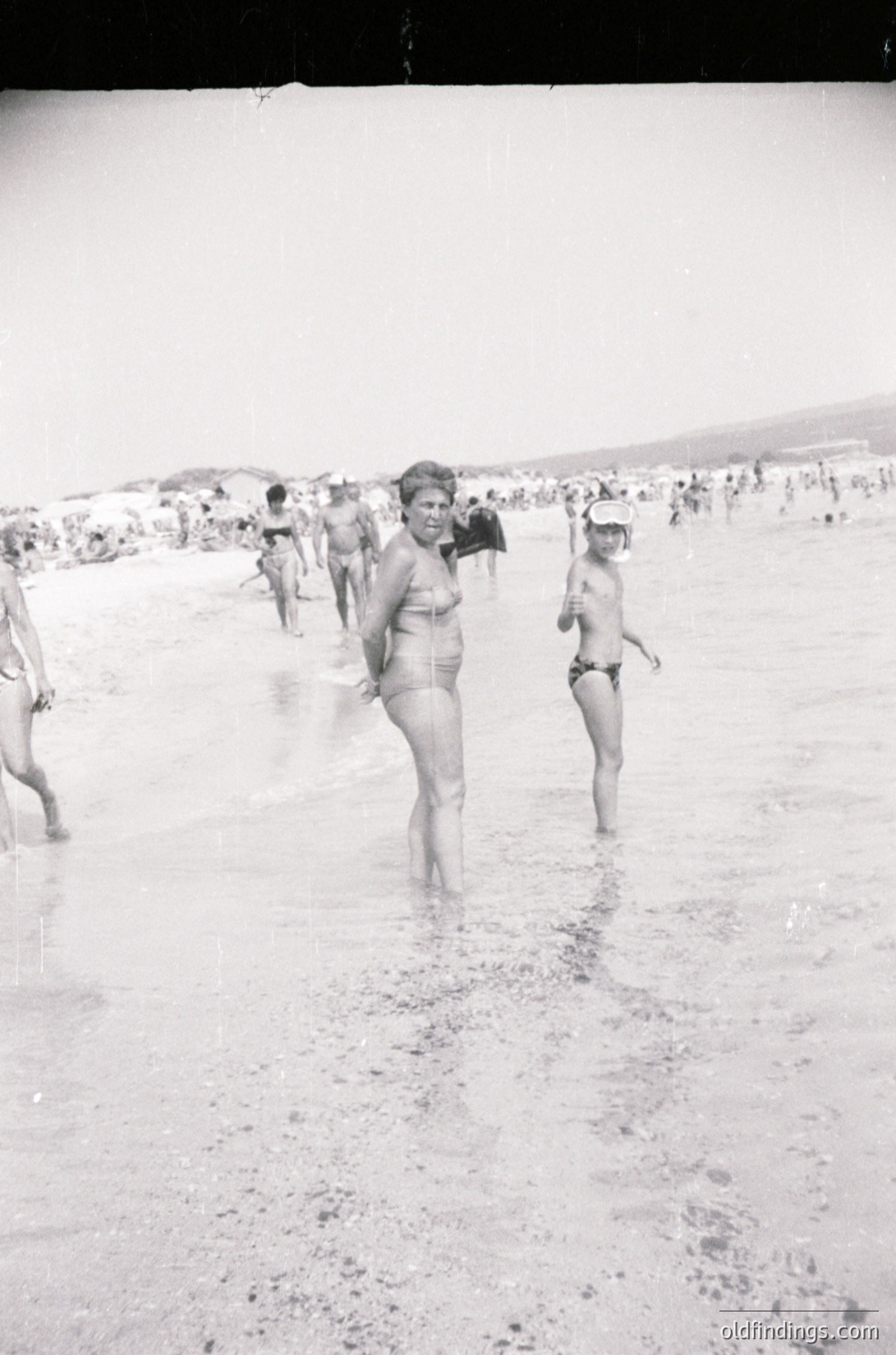 Black-and-white beach scene from the 1960s–70s, featuring mid-century swimwear and relaxed coastal culture. Central figure in a two-piece swimsuit stands on wet sand, surrounded by blurred figures in similar attire. Crowded shoreline with dunes in background.