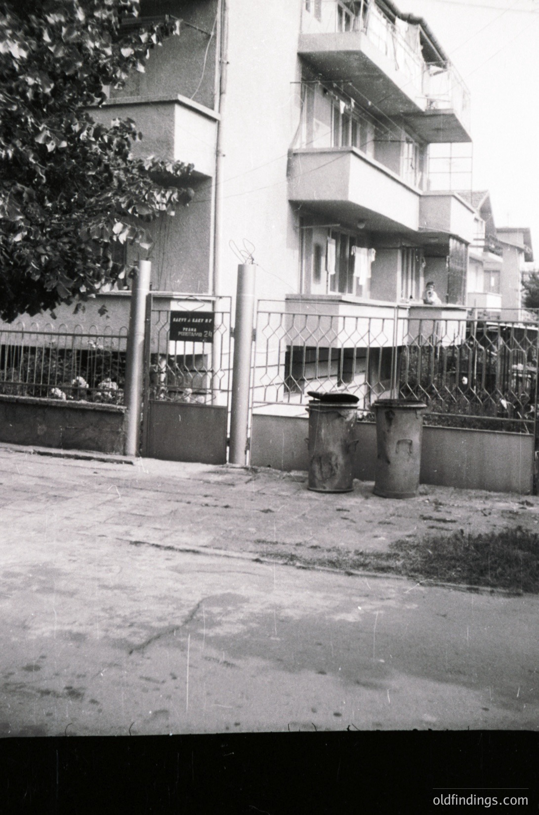 Mid-century urban courtyard with Soviet-era apartment block. Concrete balconies, barred windows, and a gated entrance sign hint at 1960s–70s Eastern Bloc architecture. Overgrown foliage and worn pavement reflect urban neglect.