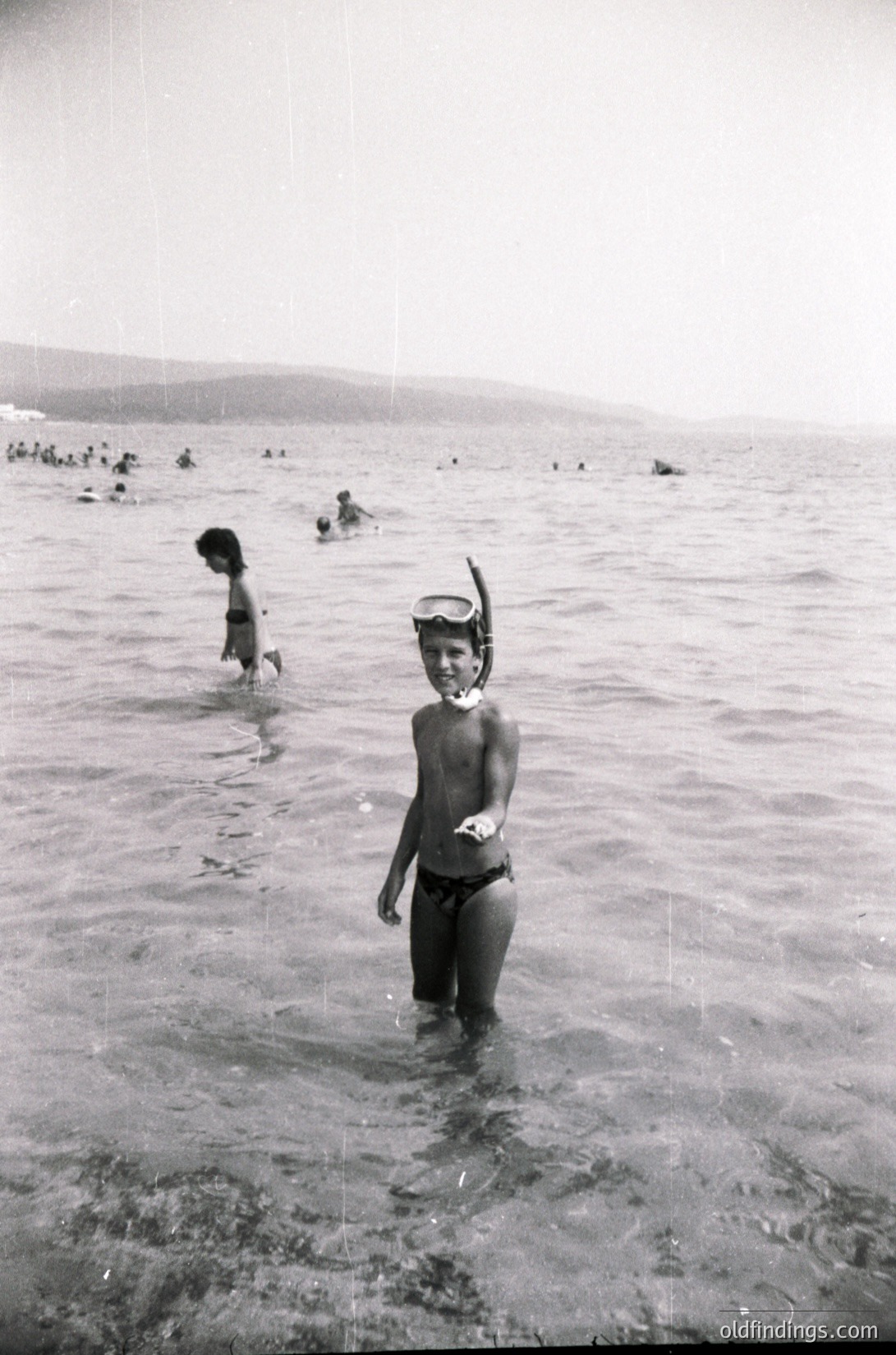 Black-and-white beach scene from mid-20th century, likely 1950s–1960s. A young boy in swim trunks and snorkel stands in shallow water, while others swim and wade in the background. Coastal landscape with distant hills and a crowded shoreline. Evokes mid-century recreational culture.
