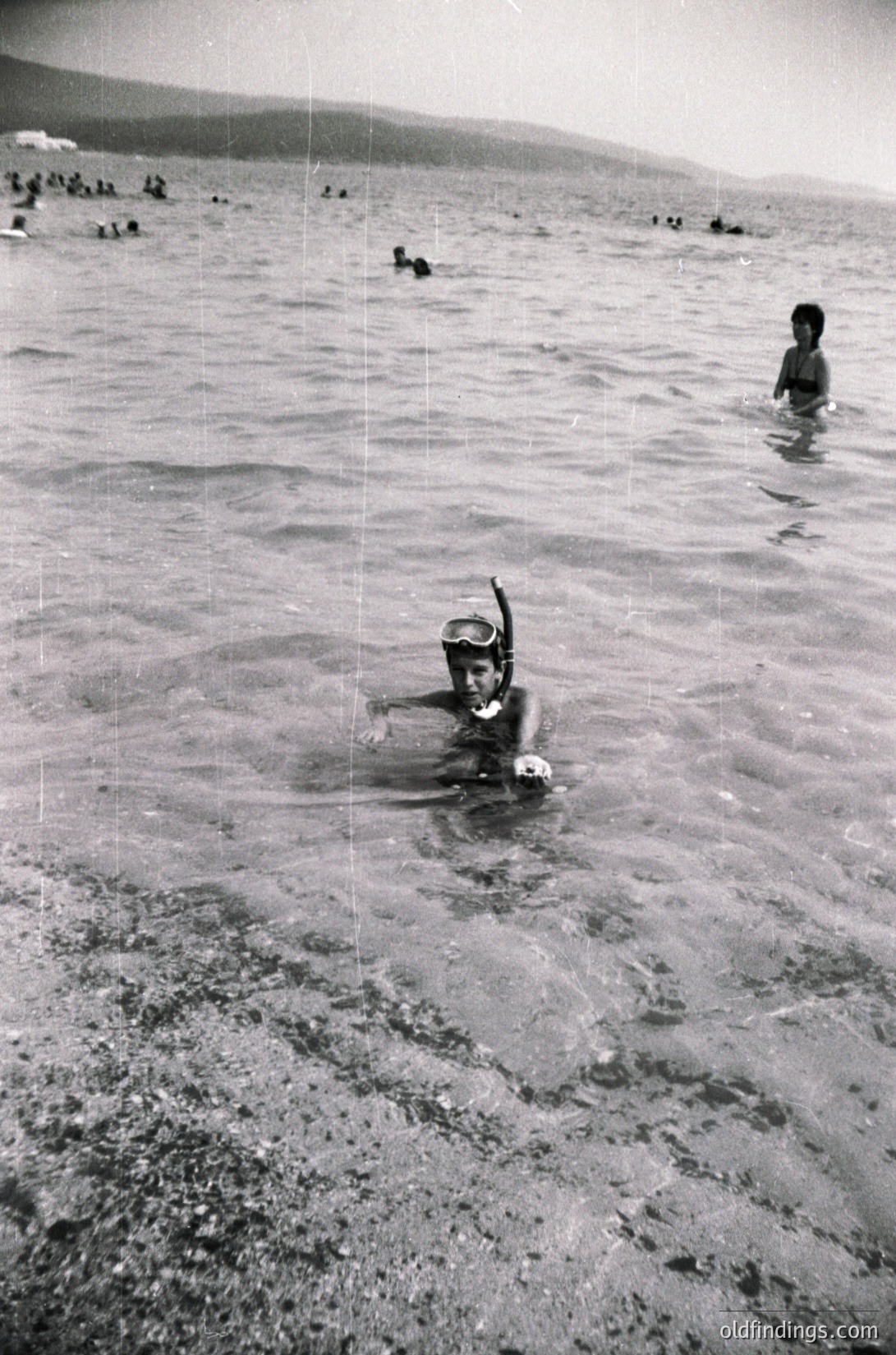Black-and-white snapshot of a mid-20th century seaside scene. A child snorkels in shallow water, wearing a snorkel mask and swim diaper, while others swim and wade in the background. Sandy beach meets calm waves under a cloudy sky. Likely a family outing or public beach during summer.
