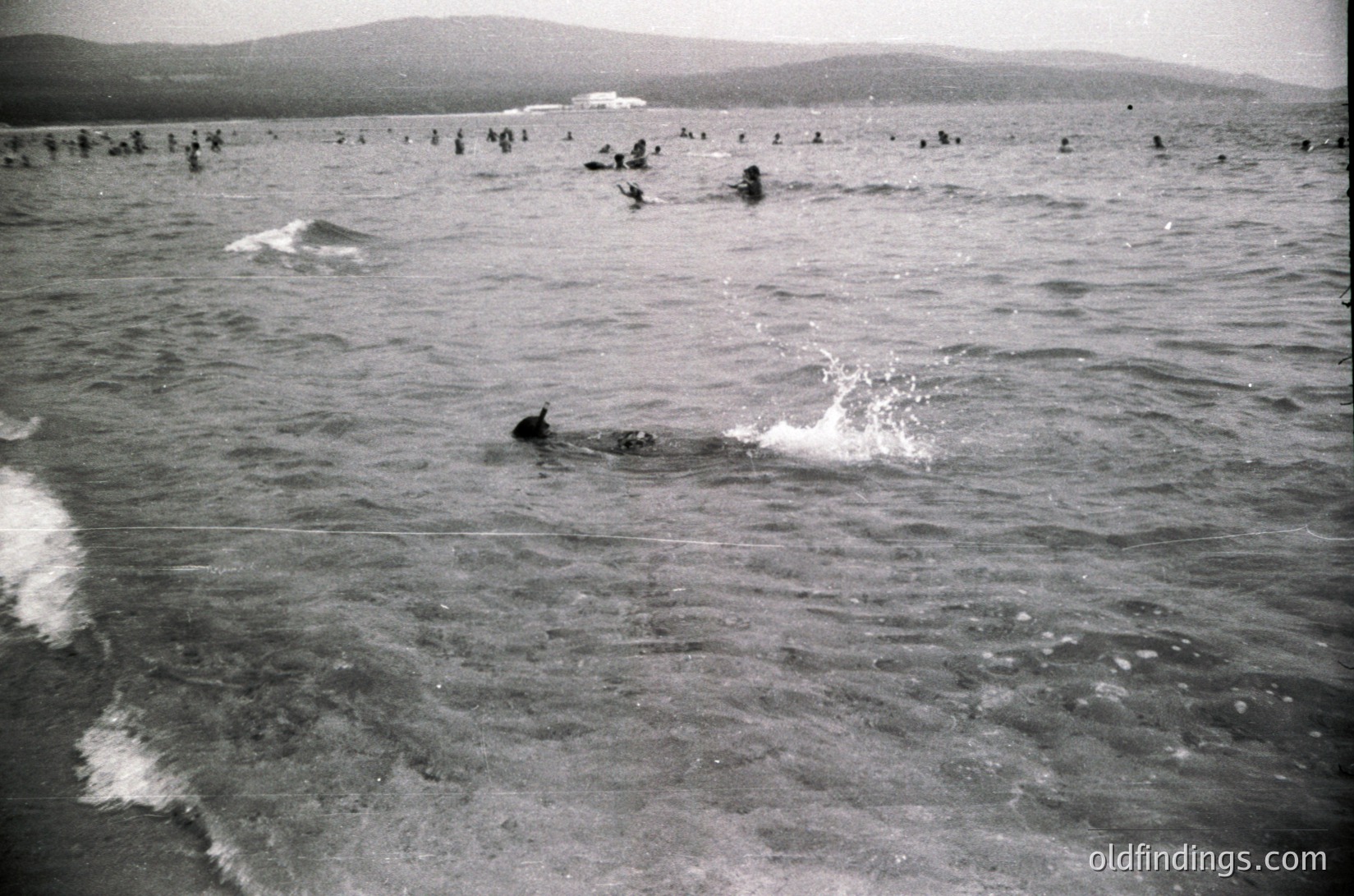 Crowded seaside scene with numerous swimmers in shallow water, likely mid-20th century. Distinctive wavy lines from a boat’s propeller in foreground. Coastal landscape with distant hills in background.