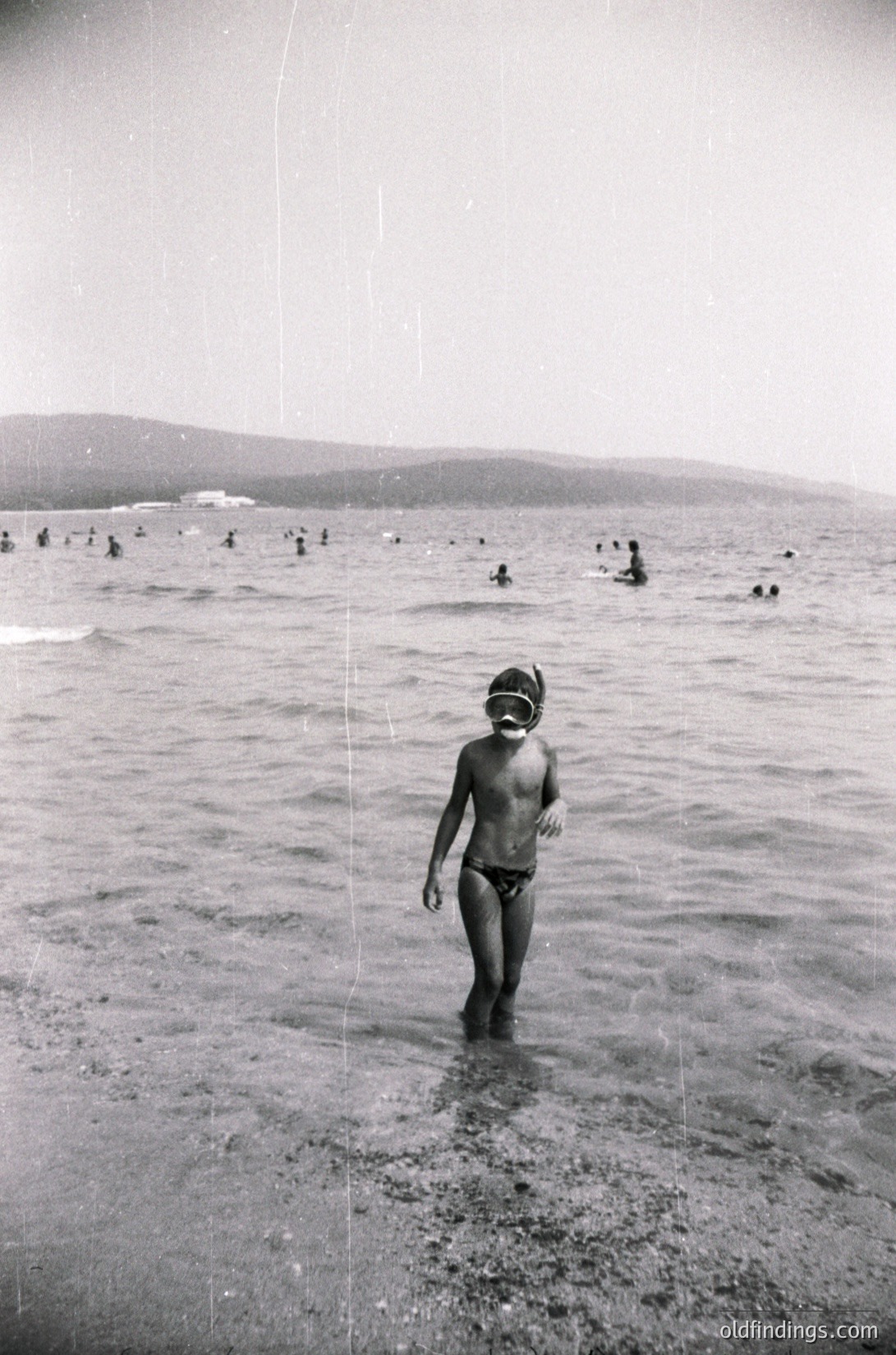 Black-and-white beach scene from the 1960s–70s, featuring a child in swim trunks and goggles wading into shallow water. Crowded shore with adults and children swimming in the background. Coastal landscape with distant hills and a hazy horizon.