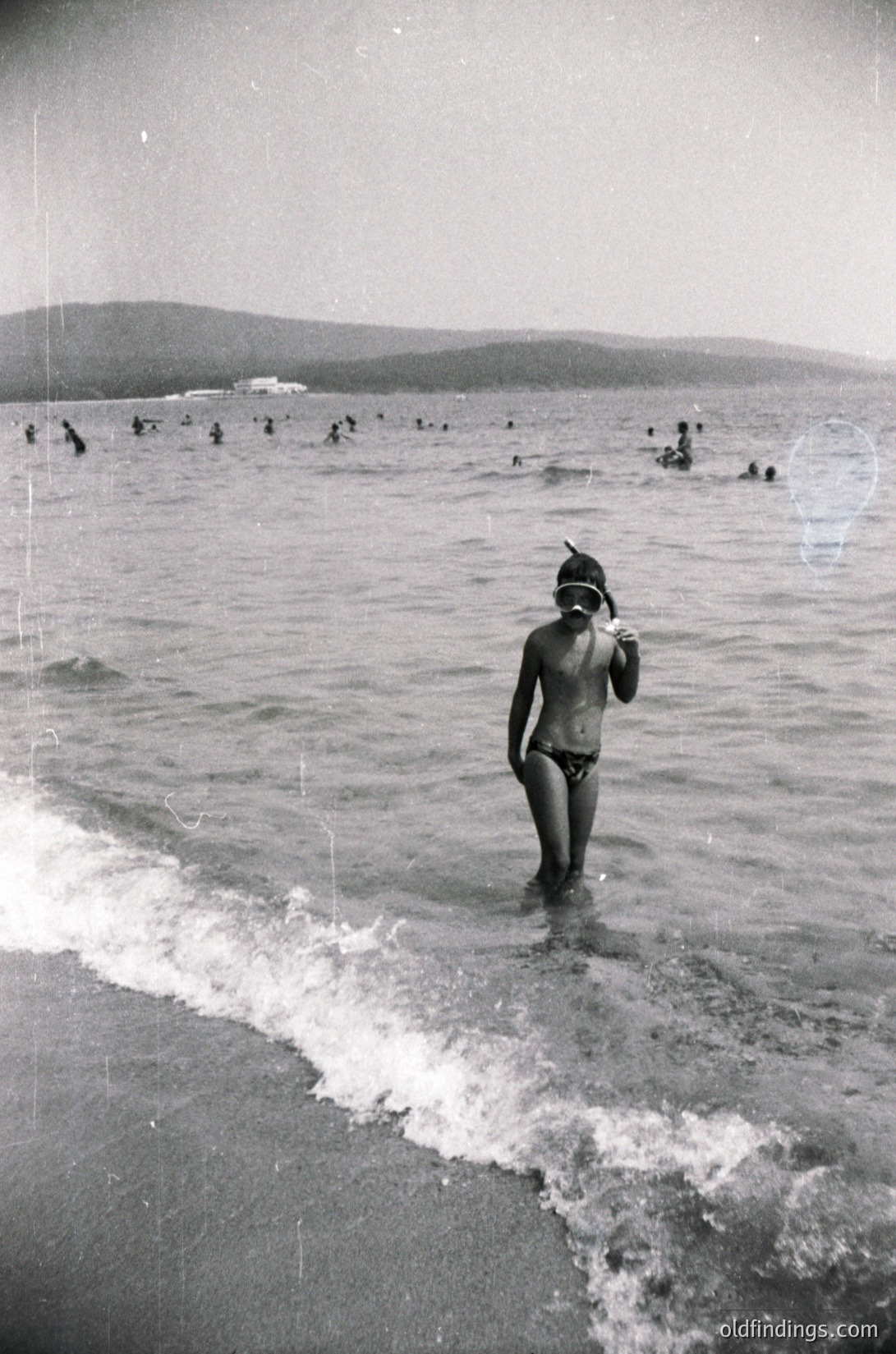 Mid-20th century seaside scene: a lone swimmer in a cap and swim trunks wades into shallow waves, while dozens of bathers dot the horizon. Coastal landscape with distant hills.
