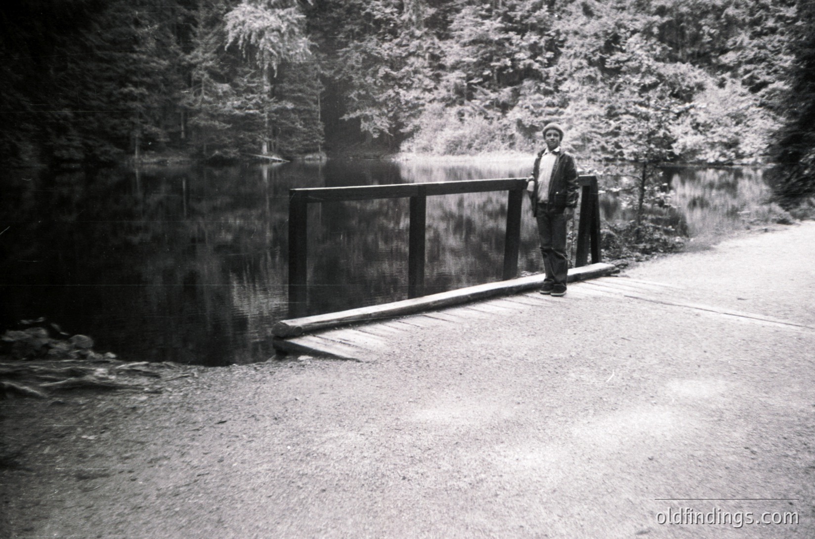 Black-and-white mid-century roadside scene featuring a man in a suit and hat standing beside a modernist metal bridge over a shallow stream. Dense forest and gravel road frame the composition, suggesting a rural or park setting. Style evokes 1950s–1960s photography.