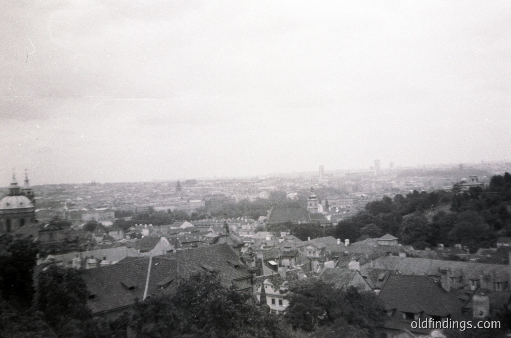 Vintage black-and-white aerial view of a European cityscape, likely early-to-mid 20th century. Dense cluster of low-rise buildings with sloped roofs, interspersed with trees and greenery. Prominent church spires and domes suggest historic architecture. Distant skyline hints at industrial or urban expansion. Foggy haze adds nostalgic atmosphere.