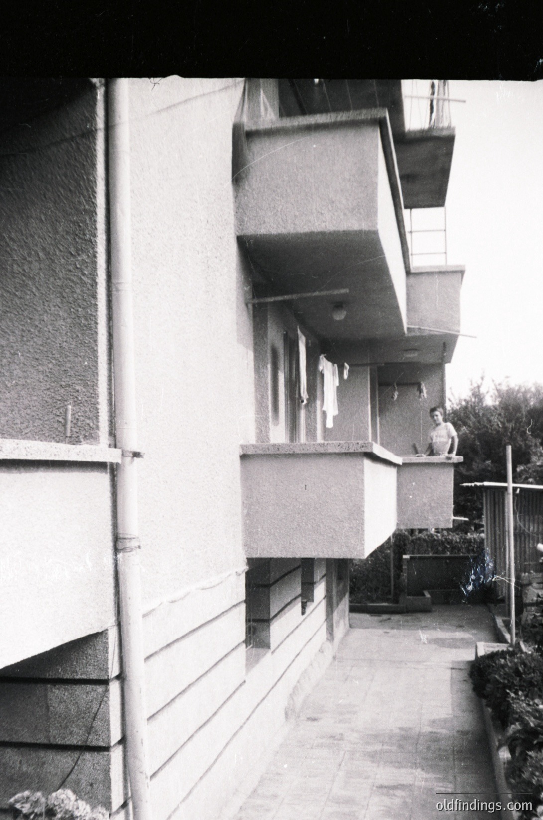 Mid-century concrete apartment balcony corridor with minimalist design—exposed pipes, flat roofs, and angled balconies. Two figures (one seated, one standing) on upper balconies, laundry hanging. Likely Eastern Bloc residential architecture, 1960s–1980s.