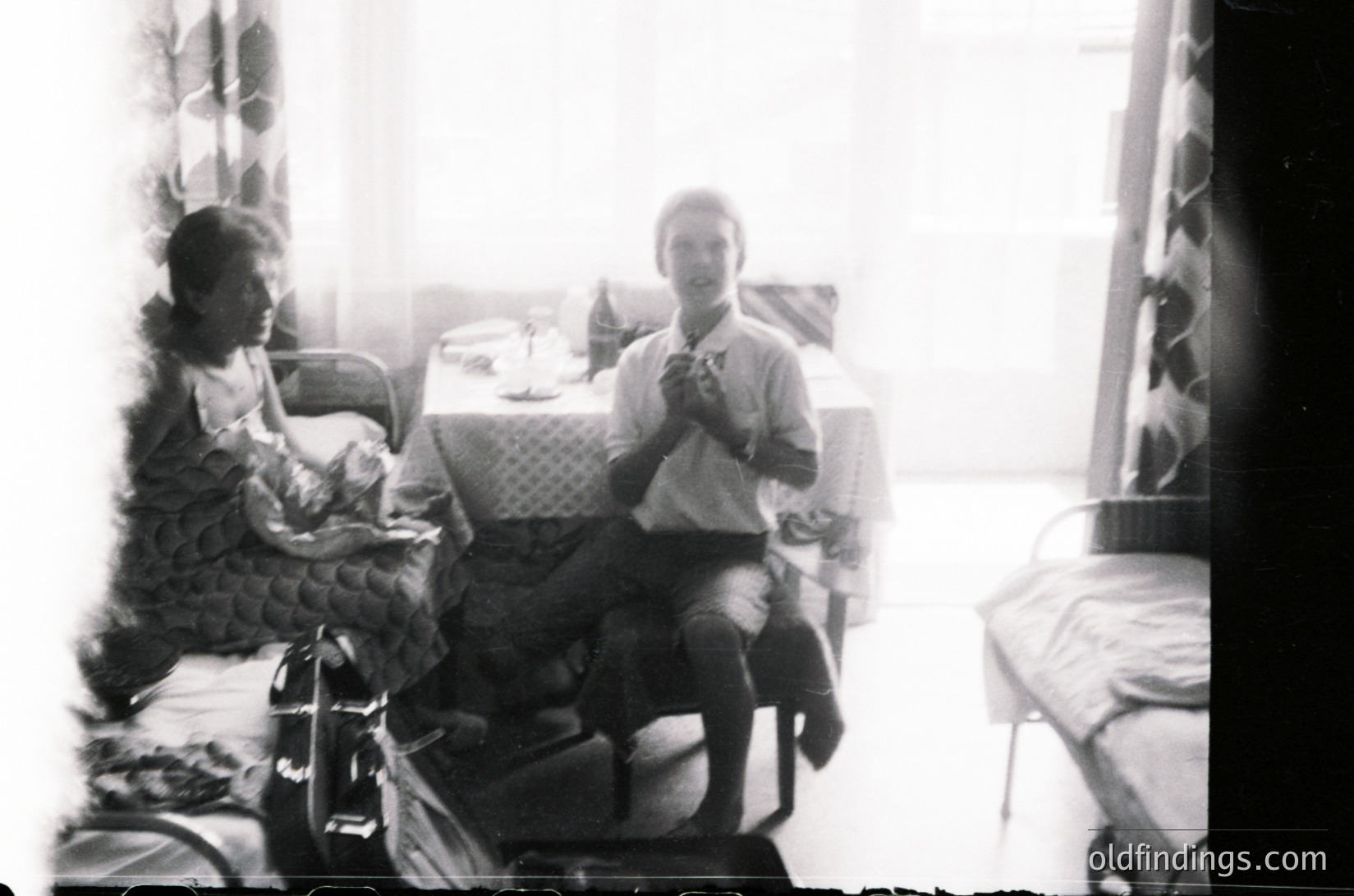 Vintage black-and-white indoor scene featuring two seated individuals in a modest room. One holds a small child, while the other clutches a cigarette. Mid-century furniture includes a woven chair, metal-framed table, and patterned wallpaper. Likely 1950s–1960s domestic setting.