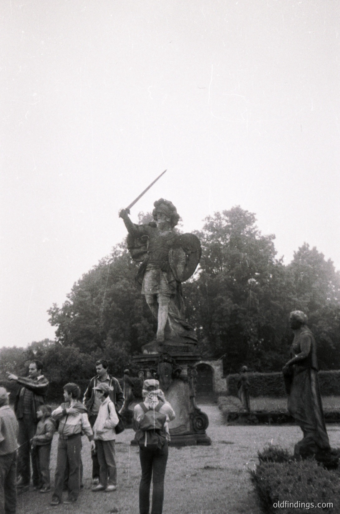 Bronze statue of a robed figure wielding a spear in a landscaped park, surrounded by a group of casually dressed children and adults. Mid-20th century outdoor setting with dense foliage and stone pathways.