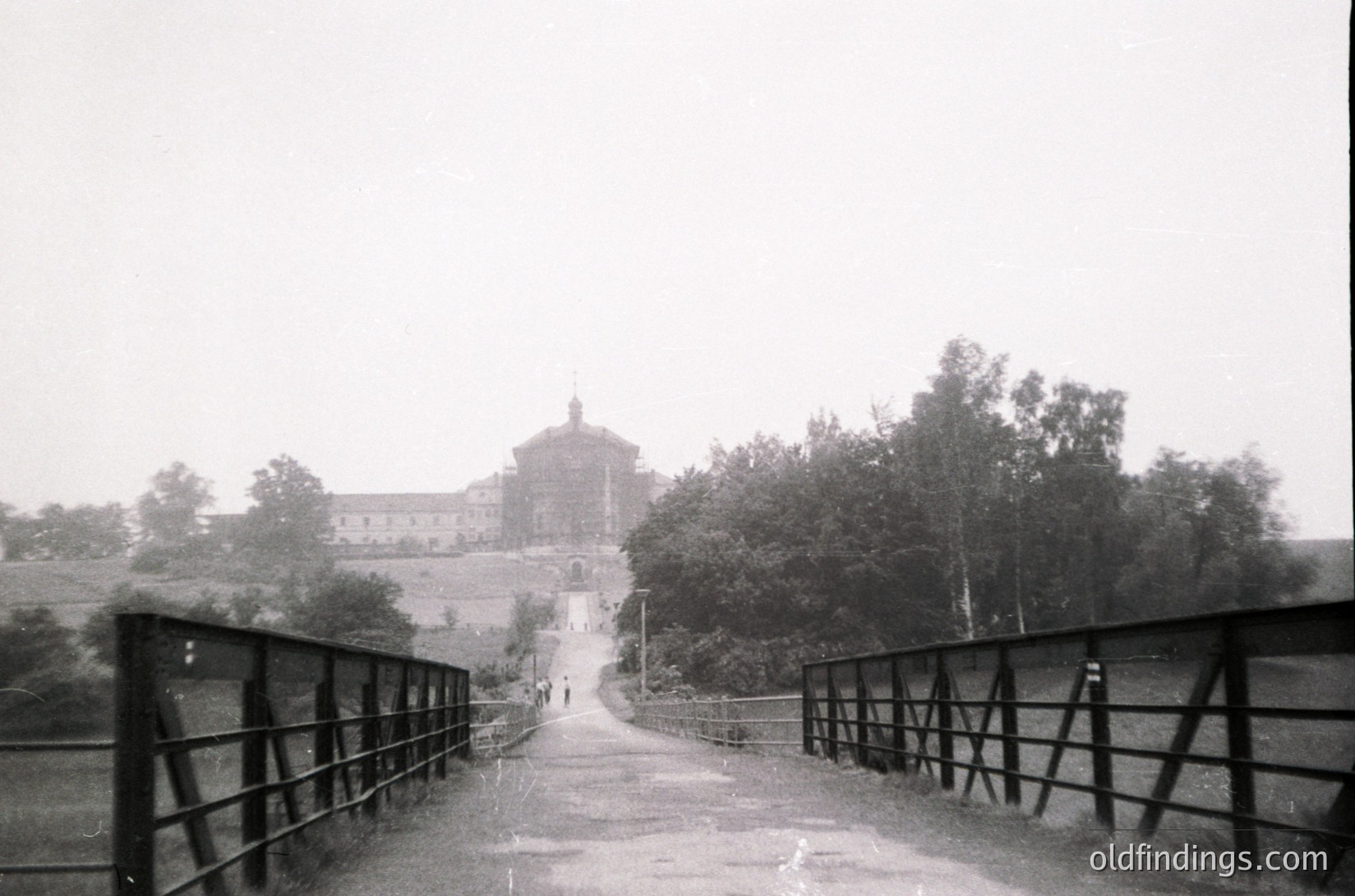Vintage black-and-white photograph of a grand, cylindrical building with a dome, likely a monastery or historic estate, framed by a metal bridge with railings. Mist or fog obscures distant details, enhancing the scene’s timeless quality. Path leads directly toward the structure, surrounded by dense greenery.