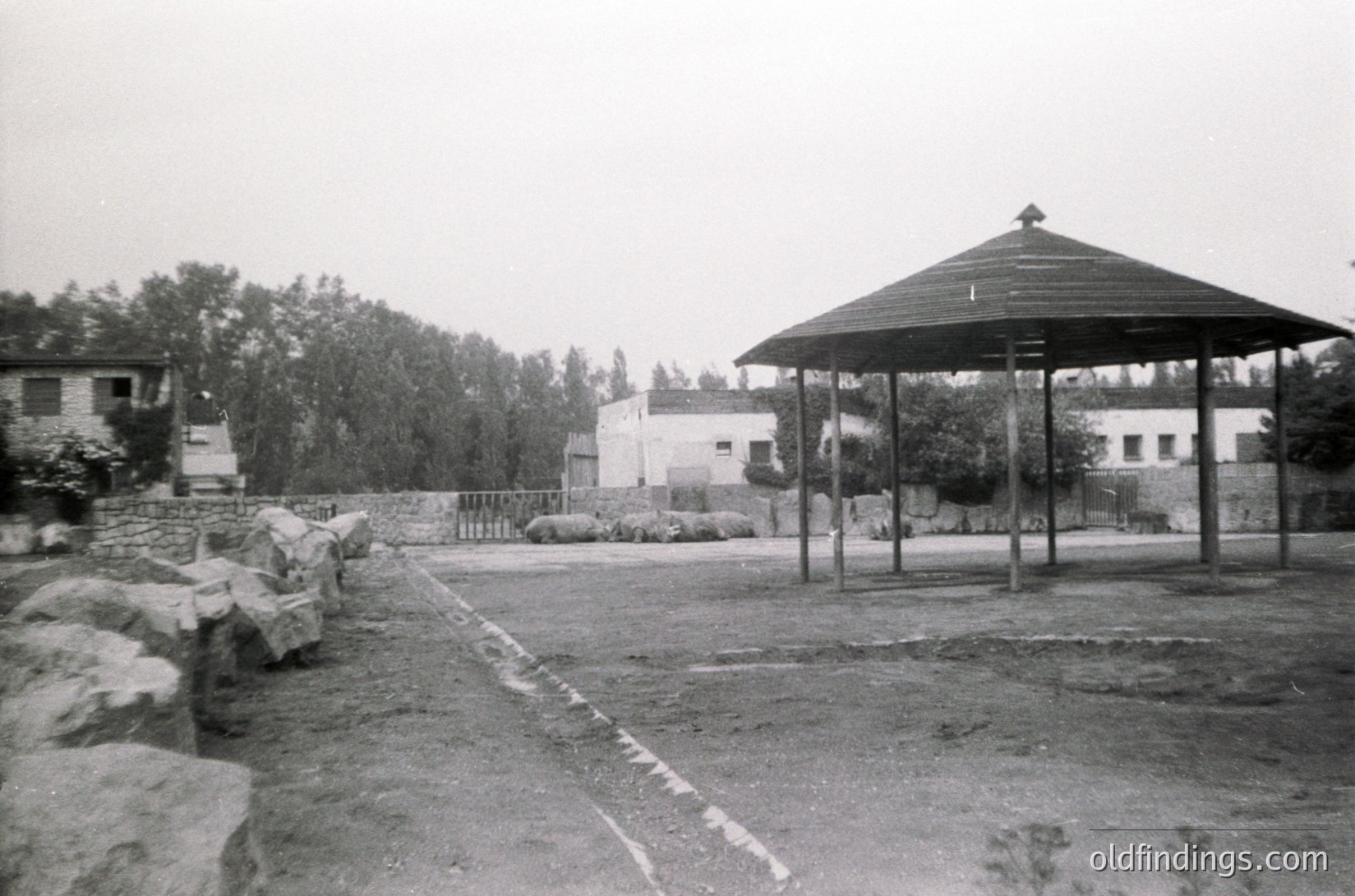 Mid-century concrete pavilion with peaked roof in a landscaped courtyard, surrounded by low stone walls and sparse trees. Residential-style buildings in background suggest institutional or public housing. Gravel pathway and scattered rocks add texture.
