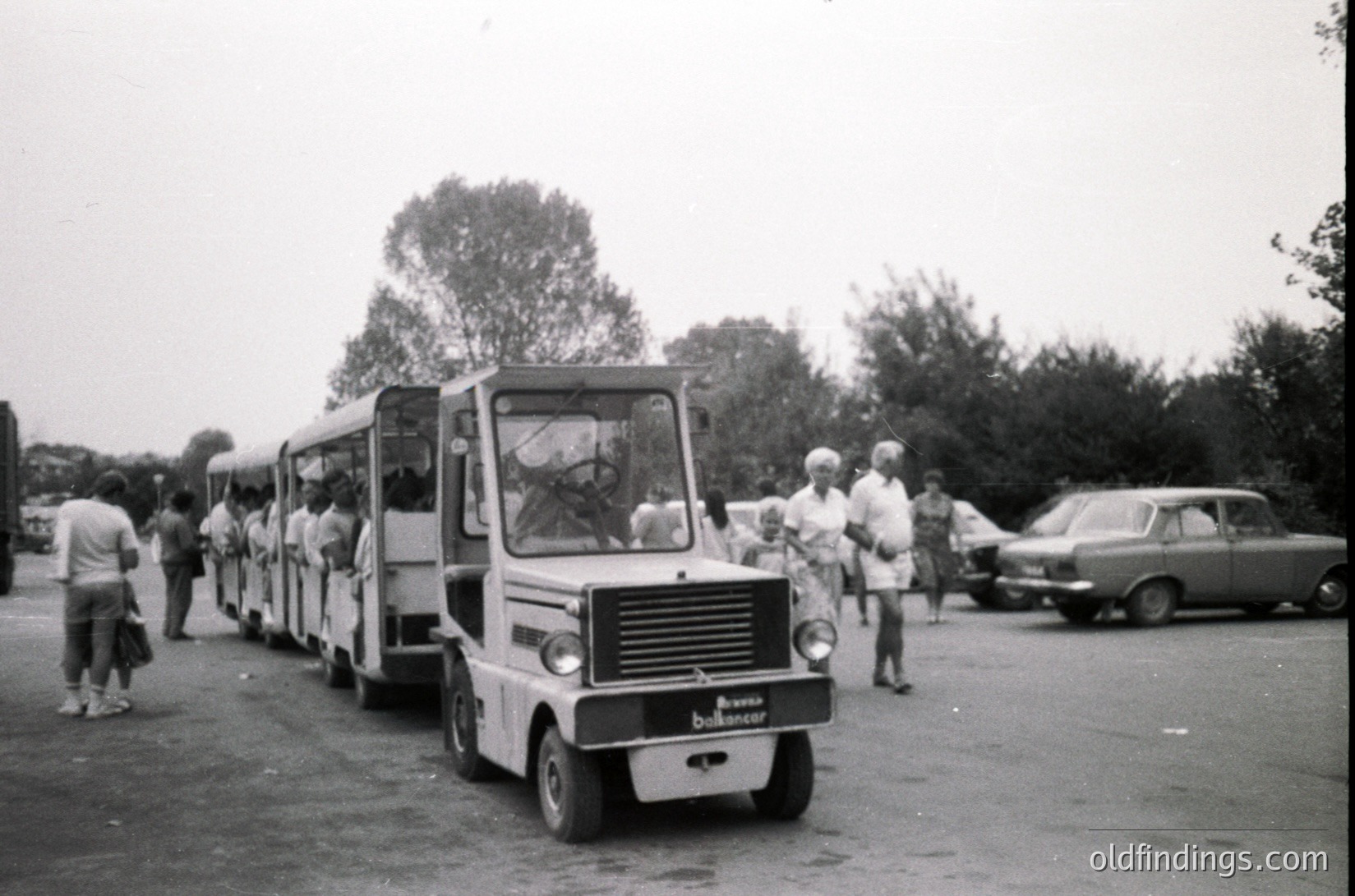 Vintage **miniature railway train** with open-air carriages, likely from the **1960s-1970s**, parked in an outdoor setting. People in retro clothing stand around, some near a classic car. The scene suggests a **theme park or amusement area**.