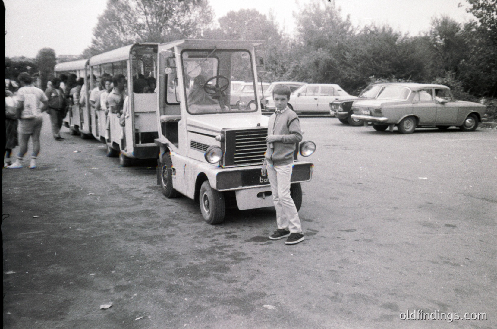 Vintage microbus parked in a lot, likely Eastern Bloc-era design. A man in 1970s-style casual wear (sneakers, baggy pants, short-sleeve shirt) poses beside it. Queue of people boarding the bus, suggesting public transport or tour service. Classic cars and trees in background indicate an outdoor urban or suburban setting.