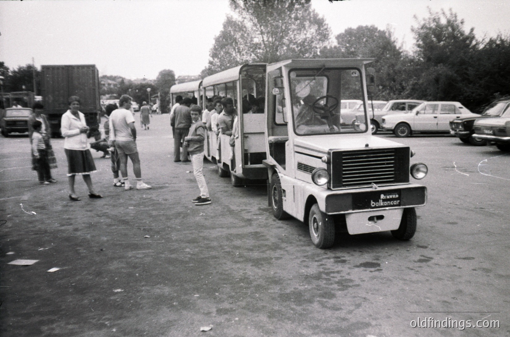 Vintage **Belfancer** shuttle bus parked in a mid-20th-century outdoor setting, likely a transport hub or industrial area. People in casual 1960s–70s attire—bell-bottoms, short-sleeve shirts—board or stand nearby. Surrounding vehicles include classic cars and a trailer truck. Bus features rounded, boxy design with "Belfancer" branding on front.