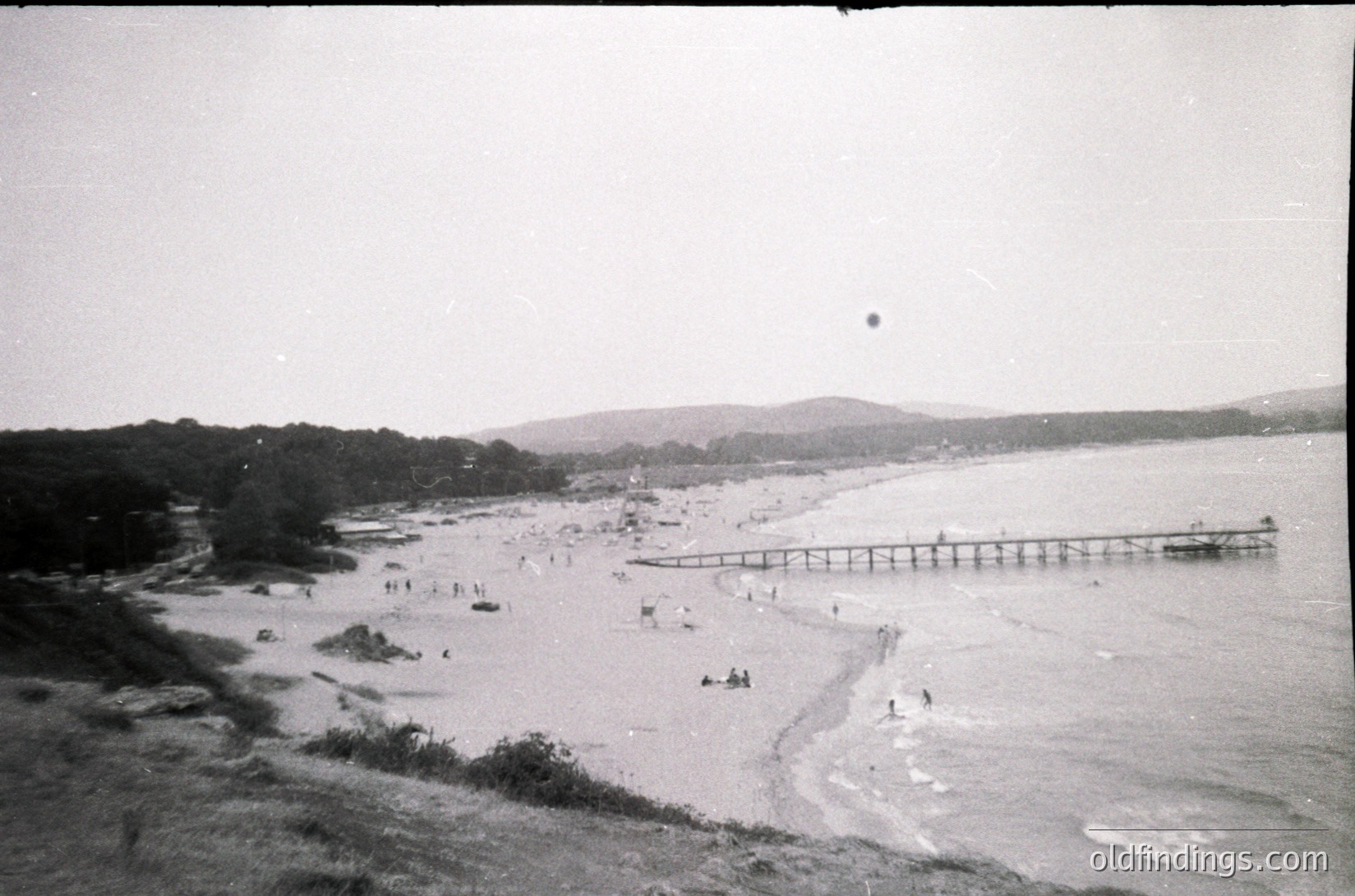 Vintage black-and-white seaside scene featuring a long pier extending into calm waters. Sandy beach with scattered groups of people sunbathing and wading. Dense pine forest on left contrasts with open coastline. Mid-20th century coastal architecture visible in background.