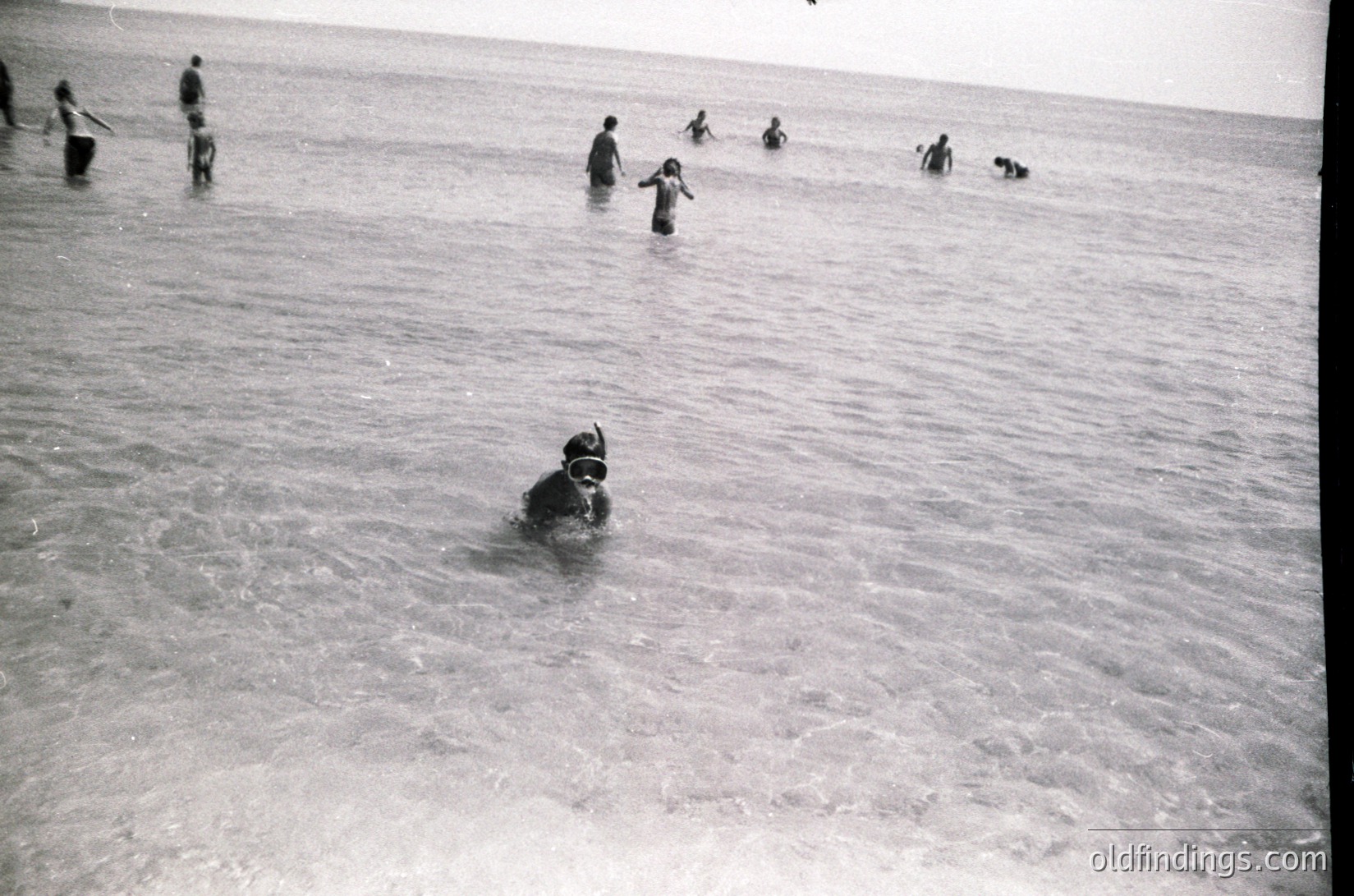 Mid-20th century beach scene with shallow waters. A scuba diver in full gear swims near the shore, while others wade or stand in knee-deep water. Casual summer attire suggests leisure activity. Likely coastal Europe or North America, 1950s-1960s.