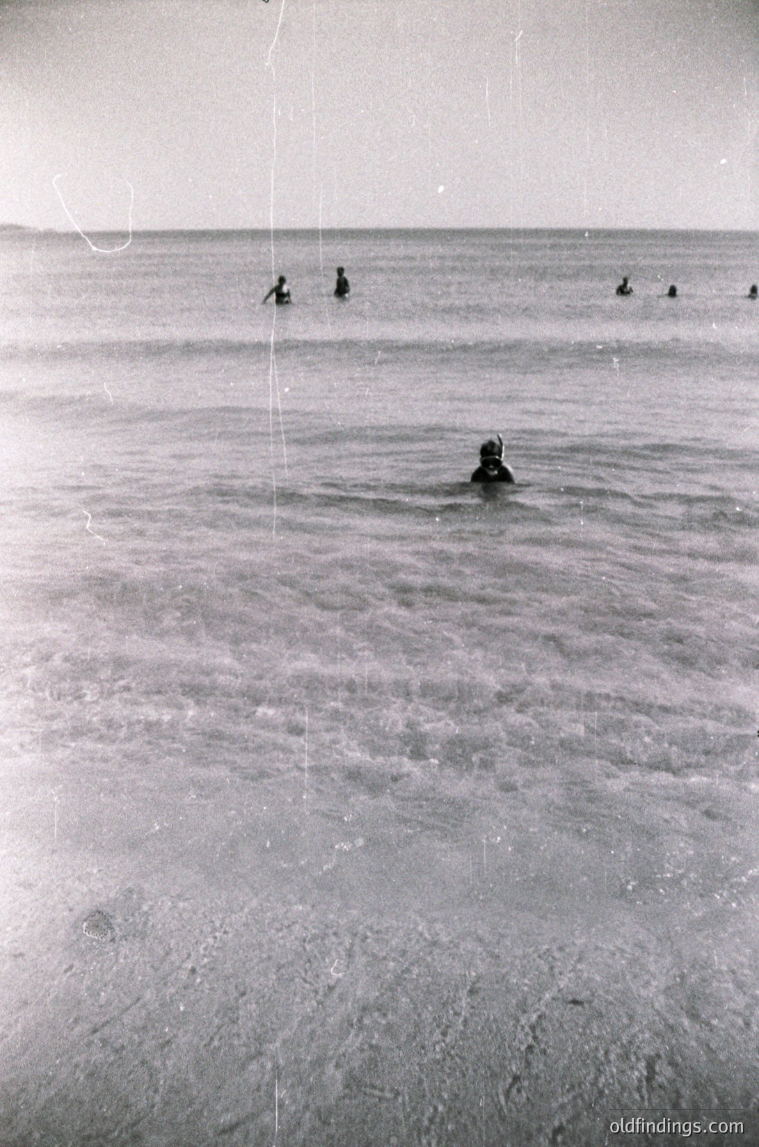 Black-and-white shot of shallow coastal waters with six figures swimming. Foreground swimmer wears a cap; background figures appear smaller, suggesting mid-20th century beach scene. Flat, sandy shore with minimal vegetation.