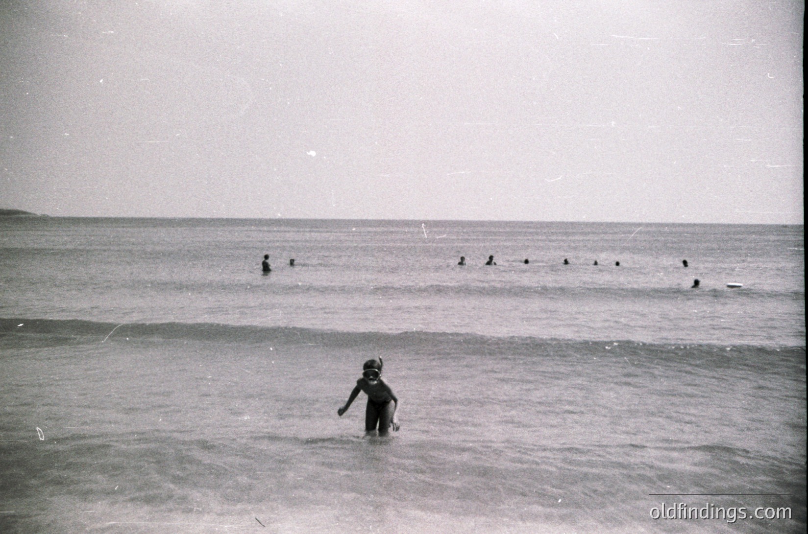 Mid-20th century black-and-white seaside scene: child wading in shallow waves, surrounded by adults swimming in deeper water. Minimalist beach attire suggests 1950s-60s era. Open horizon and calm sea evoke nostalgia for mid-century coastal leisure.