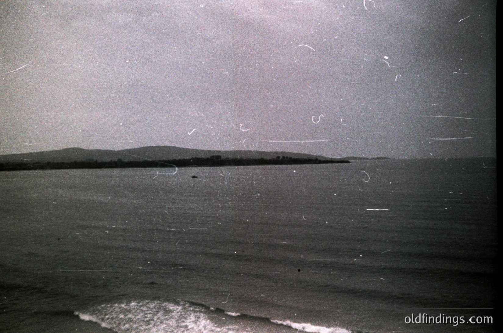 Vintage black-and-white seascape with low-contrast coastal landscape and gentle waves. Minimalist horizon line separates land and sea, hinting at a serene, possibly Mediterranean or Atlantic shoreline. Film grain and slight scratches suggest mid-20th century photography ().