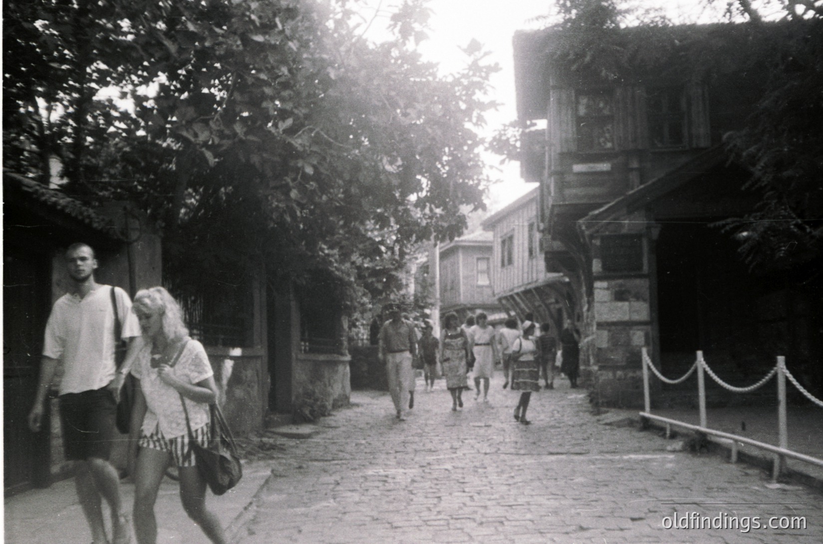 Vintage black-and-white street scene featuring cobblestone pathway flanked by wooden buildings with balconies. Mid-20th century summer attire—shorts, dresses, and hats—suggests leisurely coastal or resort atmosphere. Lush greenery and partial rope barrier indicate a pedestrian-only zone. Likely Eastern European seaside town, 1950s–1960s.