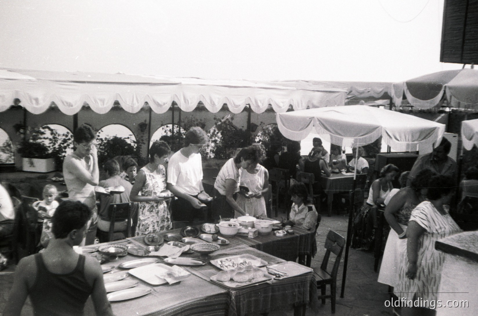 Mid-20th century outdoor café scene with white canopies and floral-patterned dresses. Tables laden with plates, cutlery, and food trays. Patrons seated and standing, engaging in conversation. Likely a seaside or resort location, suggested by relaxed attire and casual dining setup. éCulture