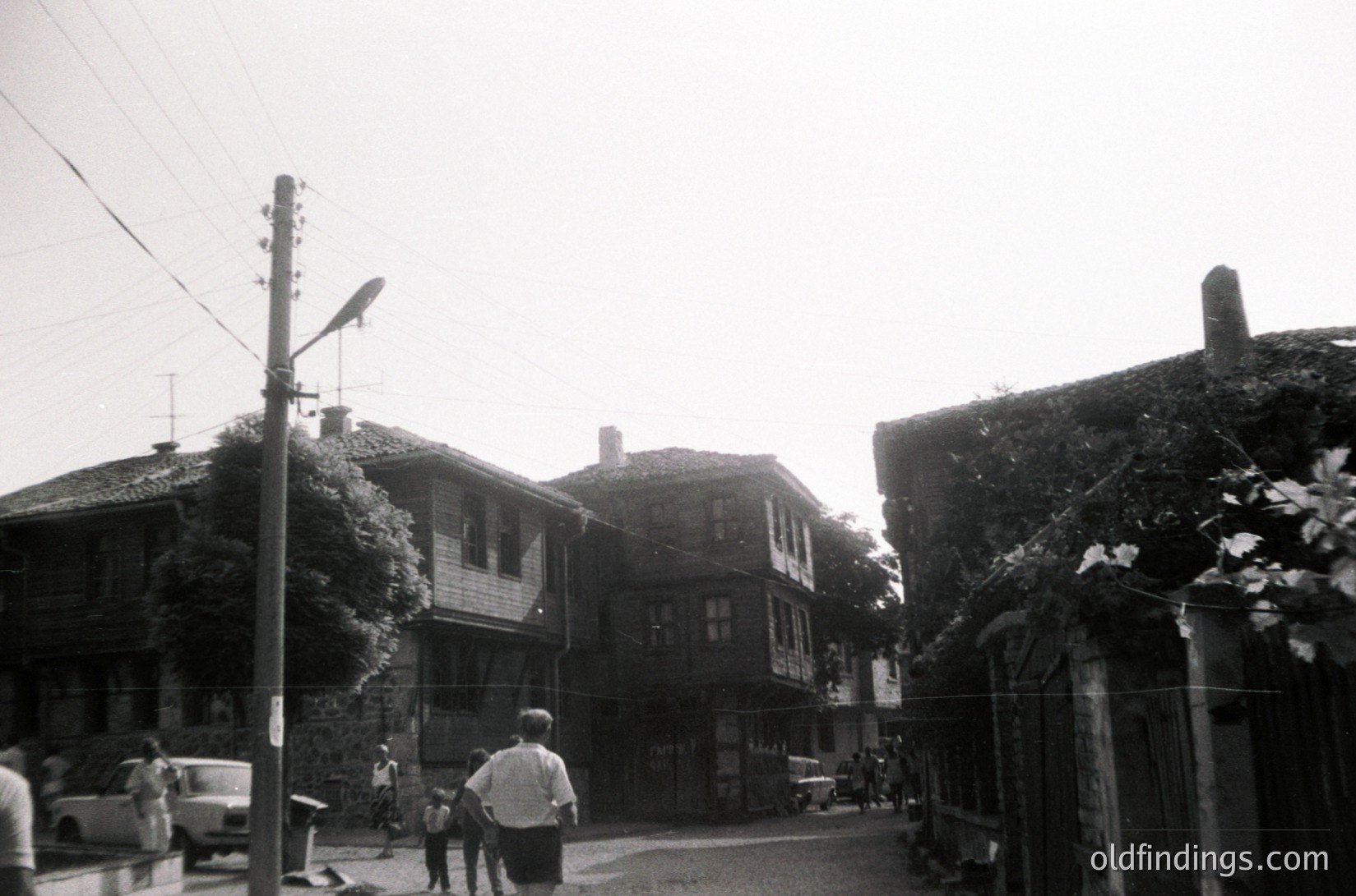 Black-and-white street scene featuring two-story wooden houses with overgrown greenery, indicative of Eastern European architecture. A man and child walk along a narrow, unpaved road flanked by vintage streetlights and utility poles. Vintage car parked on left, suggesting mid-20th century (1950s–1960s). Likely rural or small-town setting.