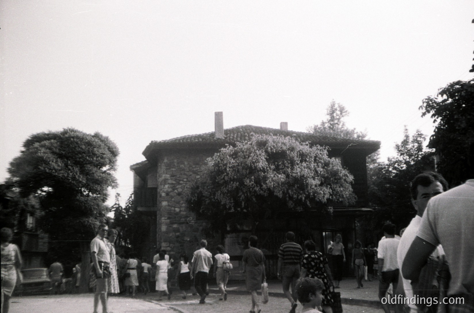 Mid-20th century courtyard scene with stone building featuring rounded corners and arched windows. Crowd of casually dressed people—men, women, and children—gathering around a central area. Lush greenery frames the building. Likely Eastern European or Soviet-era architecture.