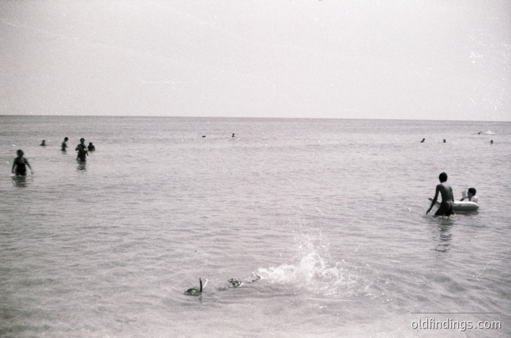 Black-and-white seaside scene with shallow waters, likely mid-20th century. Group of people wading and swimming in calm, open water; one child rides a small inflatable ring. Distant figures in the background suggest a public beach setting.