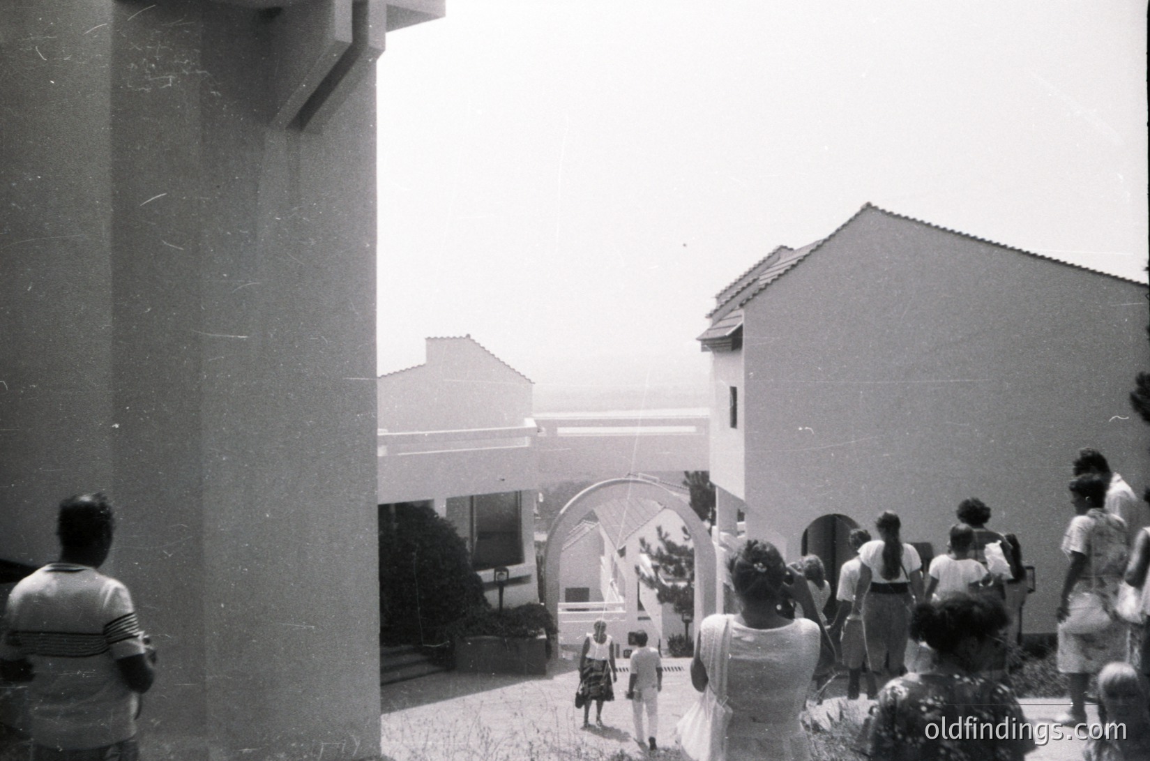 Vintage black-and-white courtyard scene with mid-century Brutalist concrete pillar framing left. Group of people in casual 1960s–70s attire (dresses, short pants, headscarves) gathered near arched entrance. Sunlit, open-air setting with minimal greenery.
