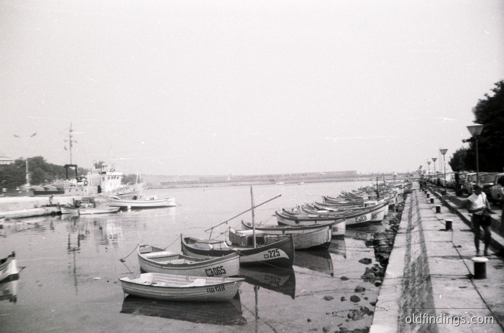 Black-and-white seaside scene featuring a row of small wooden fishing boats docked along a concrete pier. The boats display names like "Cavus" and numbered hulls (e.g., 5725). In the background, a marina with larger vessels and distant shoreline under an overcast sky. Likely –1970s Mediterranean or Black Sea port.