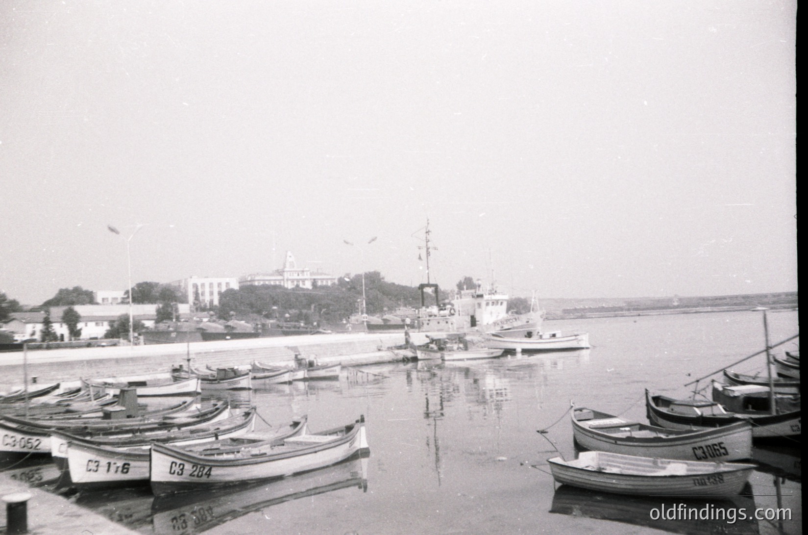 Black-and-white coastal scene featuring a marina with small wooden fishing boats marked with Cyrillic letters/numbers. Larger vessels and a prominent building with a dome in the background suggest a port town. Likely Eastern European, mid-20th century.