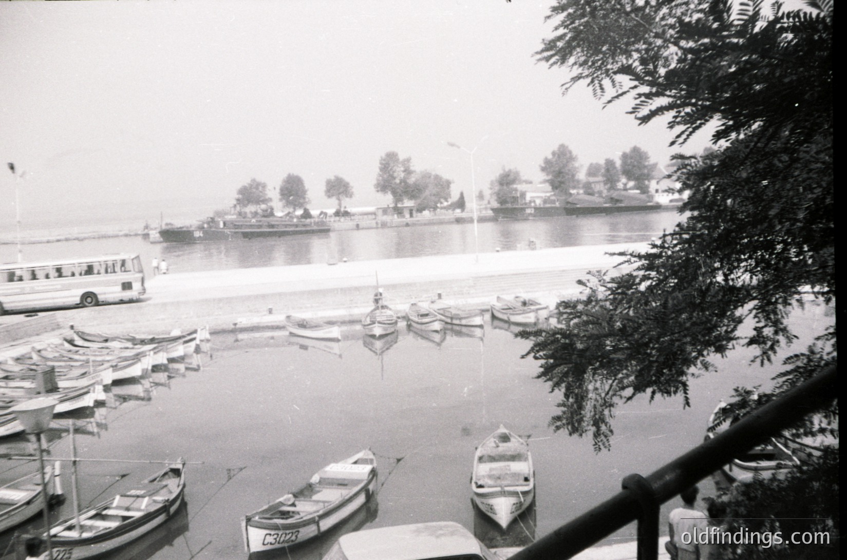 Black-and-white harbor scene featuring small wooden boats docked along a calm waterfront. A mid-20th century passenger ferry (likely 1950s–1970s) and industrial barge in background. Dense foliage frames right edge; distant trees and buildings suggest urban coastal setting.