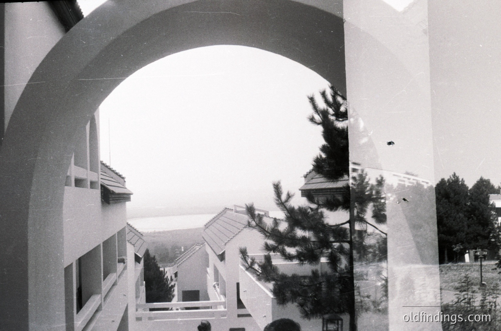 Mid-century Brutalist courtyard framed by a large archway, showcasing raw concrete structures and sloped roofs. Overlaid transparency reveals a second perspective, blending architectural layers. Likely Eastern European institutional or residential complex, 1960s–1970s.