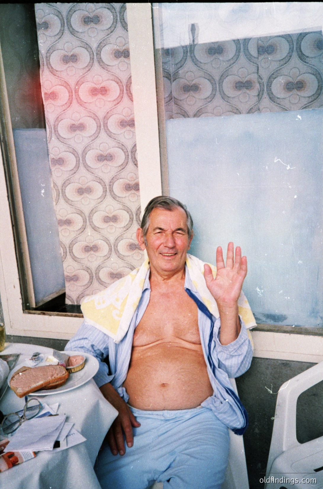 Mid-century hotel interior featuring floral-patterned wallpaper and worn furnishings. Man in loose, light-colored pajama-style attire waves toward camera, seated at a small table with a half-eaten sandwich. Sunlight streams through a partially open window, illuminating the scene. Likely 1960s–1970s seaside or resort setting.