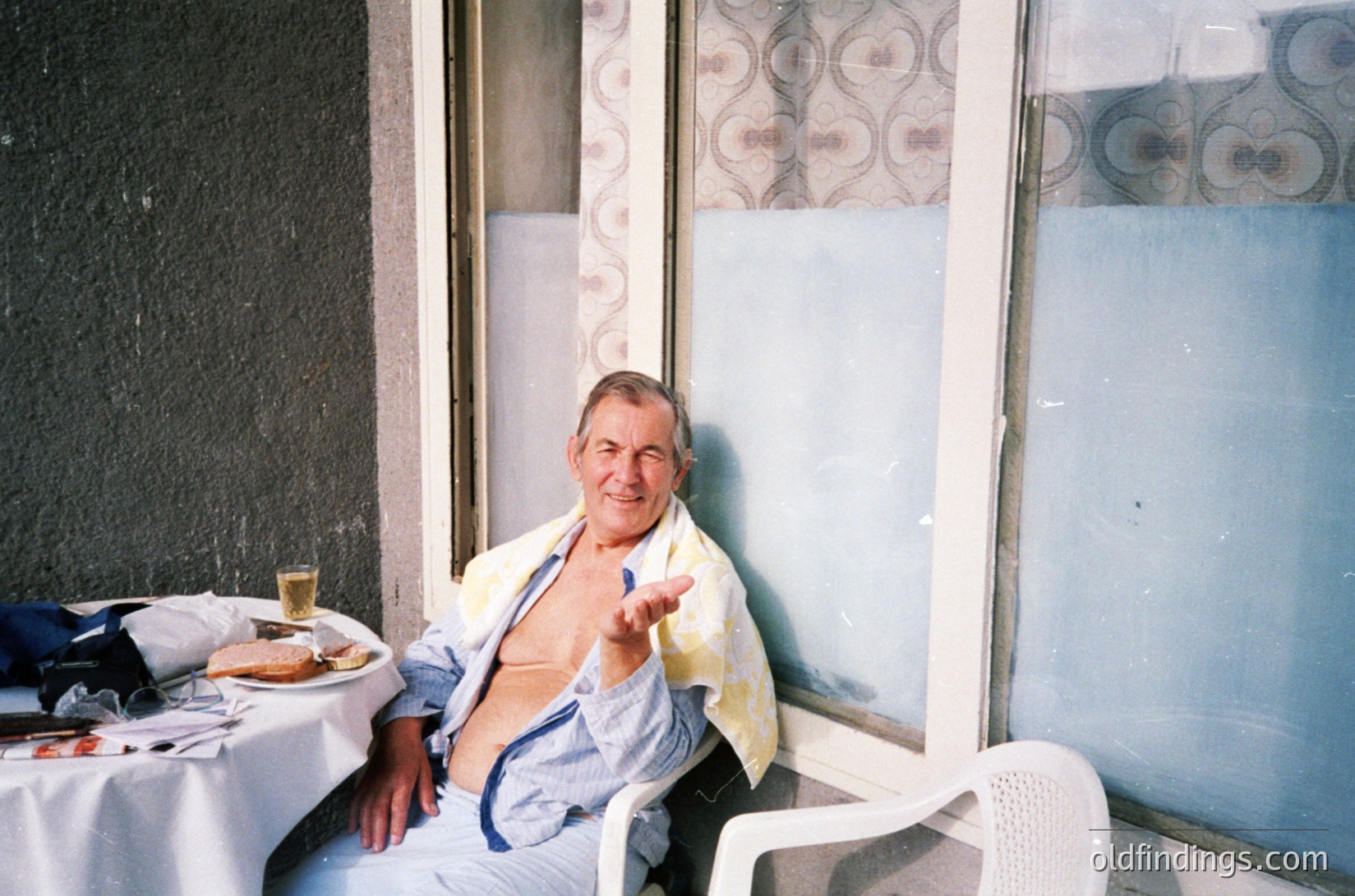 Mid-century man poses indoors with a relaxed gesture, seated in a white plastic chair. A table beside him holds a half-eaten meal and a drink. Decor features geometric wallpaper and frosted glass doors. Likely a café or restaurant setting, suggesting a casual, social atmosphere. éCulture