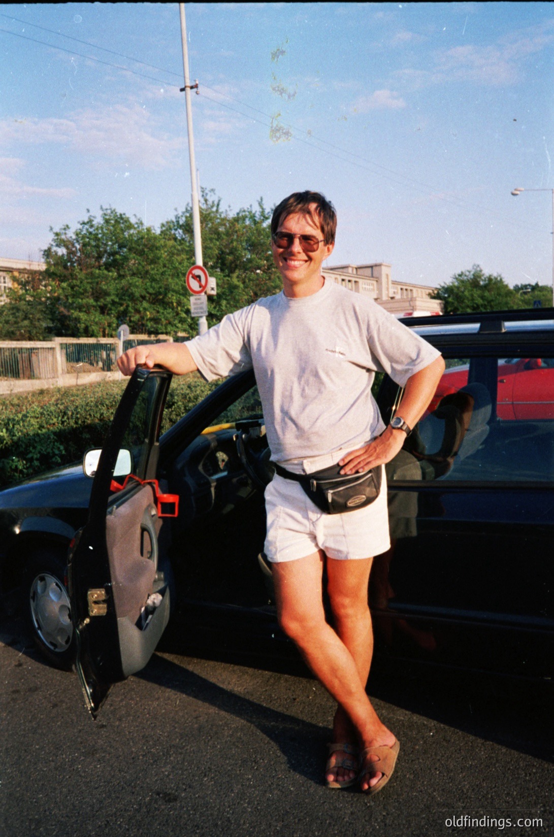 Casual 1990s street portrait: individual in white athletic wear (shirt, shorts, flip-flops) poses by a vintage hatchback car, wearing a fanny pack and sunglasses. Urban setting with a "no parking" sign and mid-rise buildings in background.