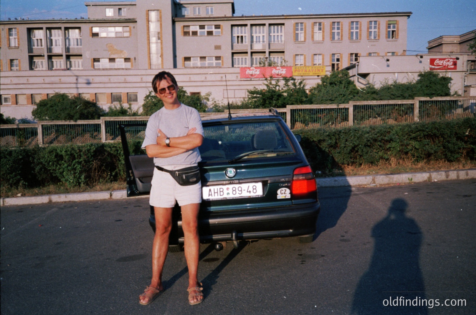 Vintage 1980s-90s urban portrait: woman in retro sunglasses, white top, and shorts poses beside a dark sedan (BMW 5-series) with Bulgarian license plate (AWB89-48). Multi-story concrete apartment block with faded Coca-Cola ads in background. Street scene captures midday sunlight and urban decay.