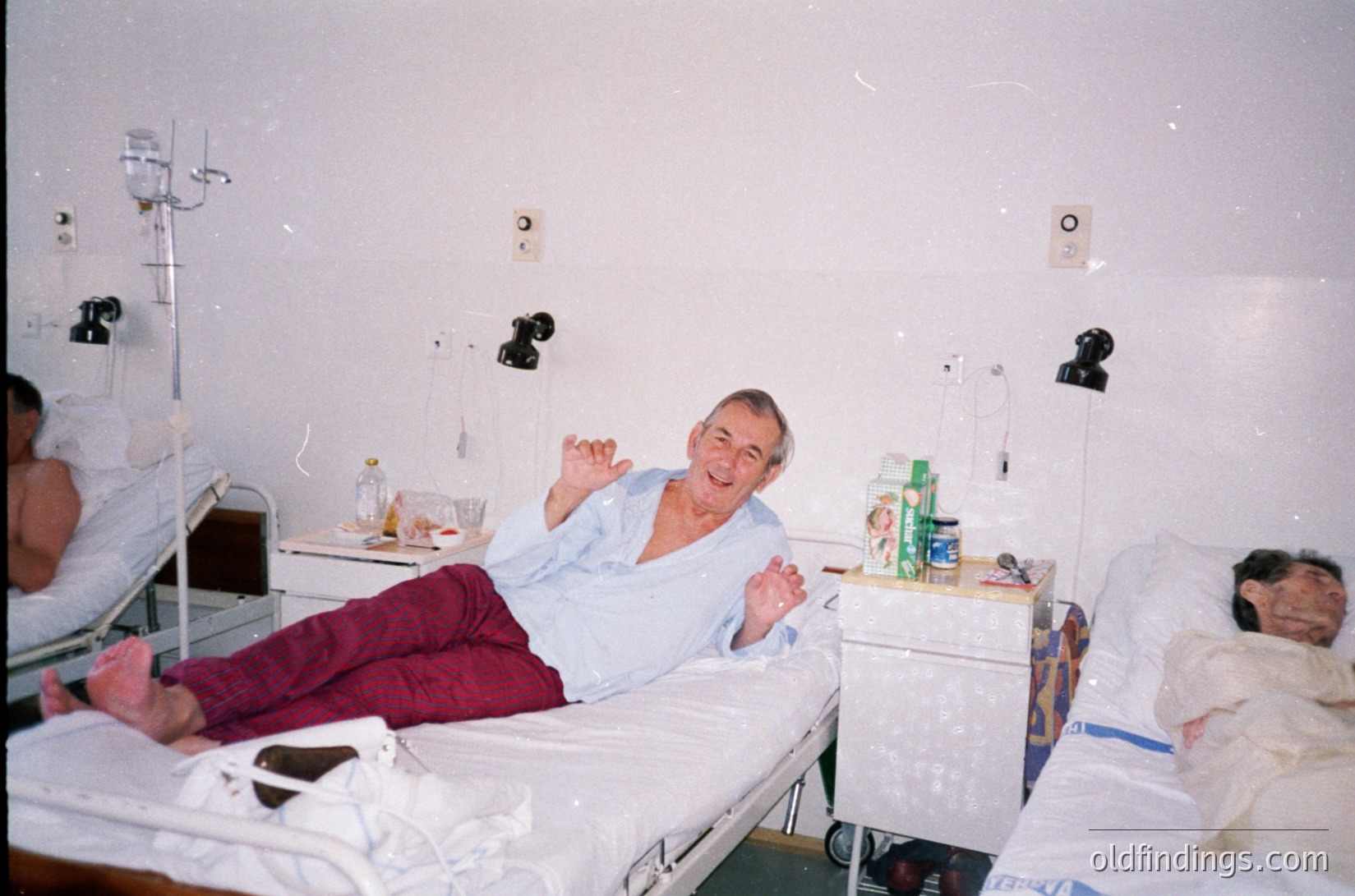 A hospital ward from the 1990s–2000s, featuring three patients in shared beds. Central figure reclines in a light blue hospital gown, smiling and waving, with medical equipment (IV poles, bottles) visible. Adjacent beds include a partially visible patient on the left and another with a blanket on the right. Bottled water and a small plastic container on the bedside table. Institutional lighting and white walls emphasize clinical setting.