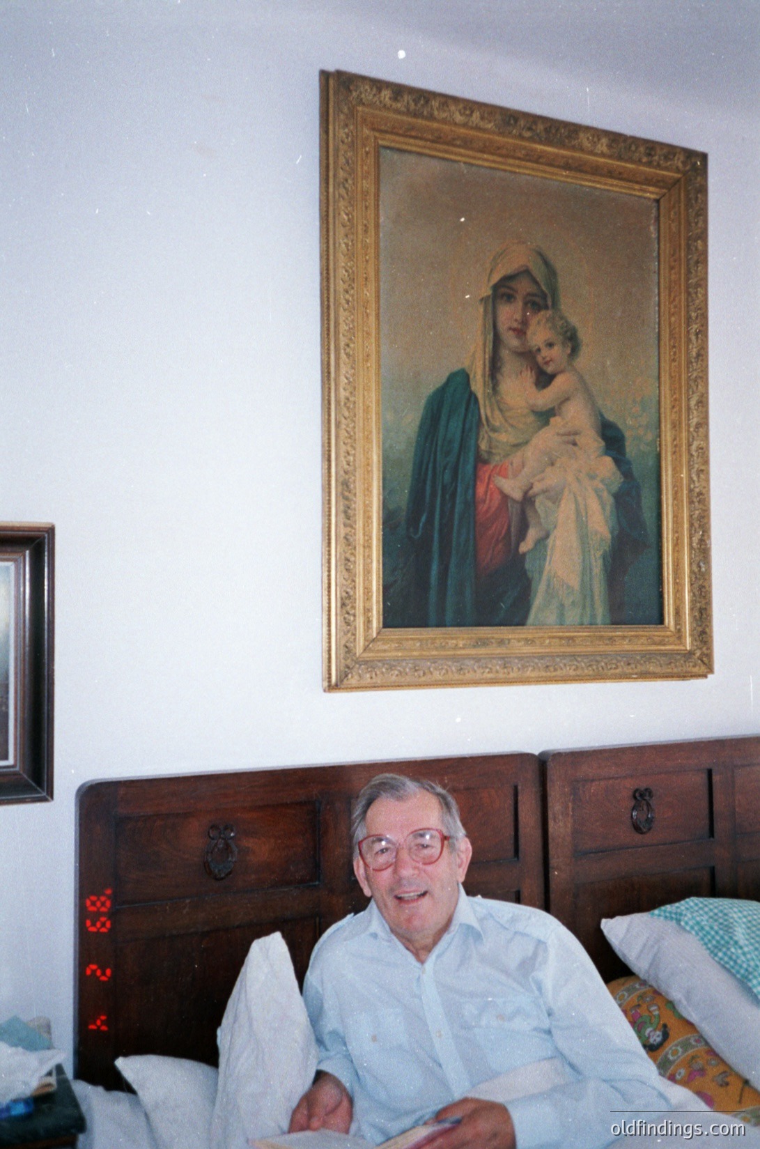 Classic religious portrait of Madonna and Child in ornate gilded frame, likely 19th–early 20th century European. Elderly man in vintage glasses and light-colored shirt poses beside a wooden headboard with carved knobs. Interior suggests mid-20th century domestic setting.