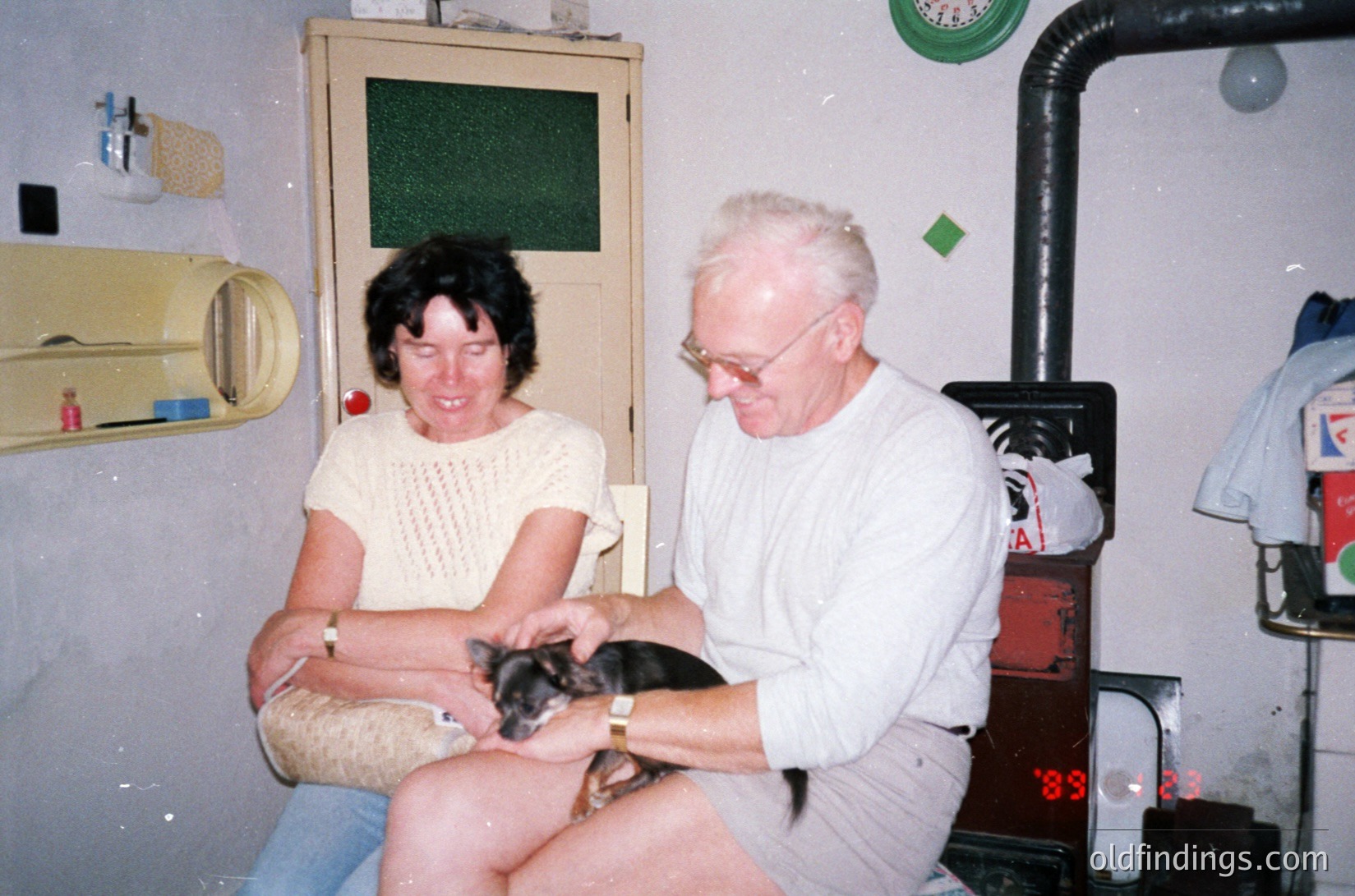 A mid-20th-century indoor scene featuring an elderly couple bonding with a small black-and-tan dog. The man, wearing glasses and a white sweater, holds the dog while the woman, in a light sweater and jeans, smiles. Background includes a vintage stove with a visible "89" serial number, a green-framed mirror, and a wall-mounted shelf with a yellow container. Likely residential kitchen or living area, 1980s–1990s.
